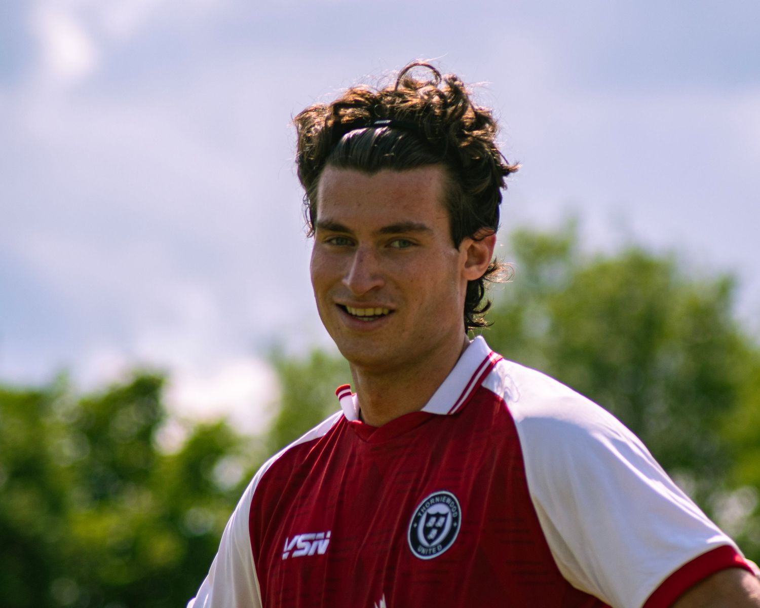 Young male soccer player in red and white jersey smiling on outdoor field with green trees in background.