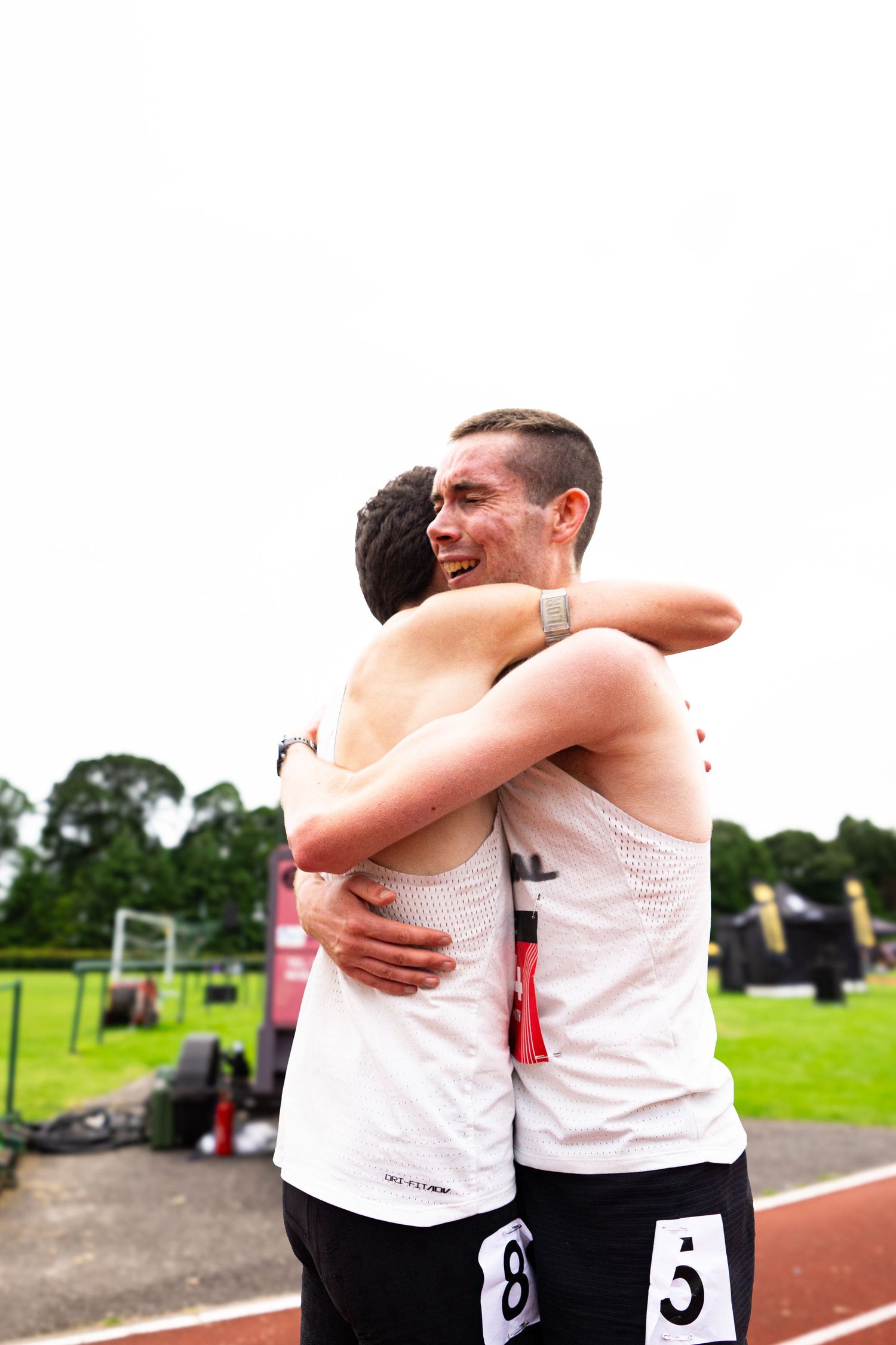 Two male athletes in white jerseys embrace on a running track after a race, smiling in celebration.
