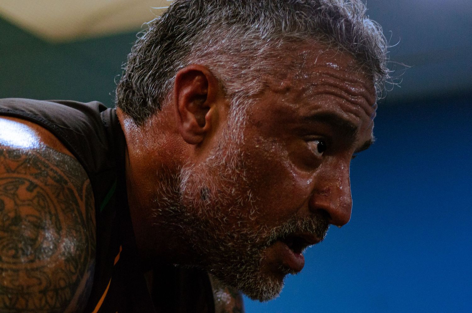 Close-up profile of a middle-aged man with gray stubble and a shoulder tattoo, looking downward intensely.