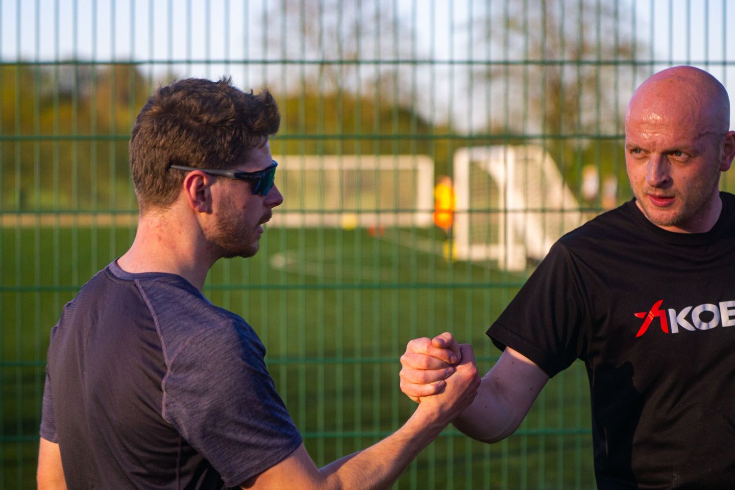 Two male runners shake hands in sportsmanship near a chain-link fence after training.