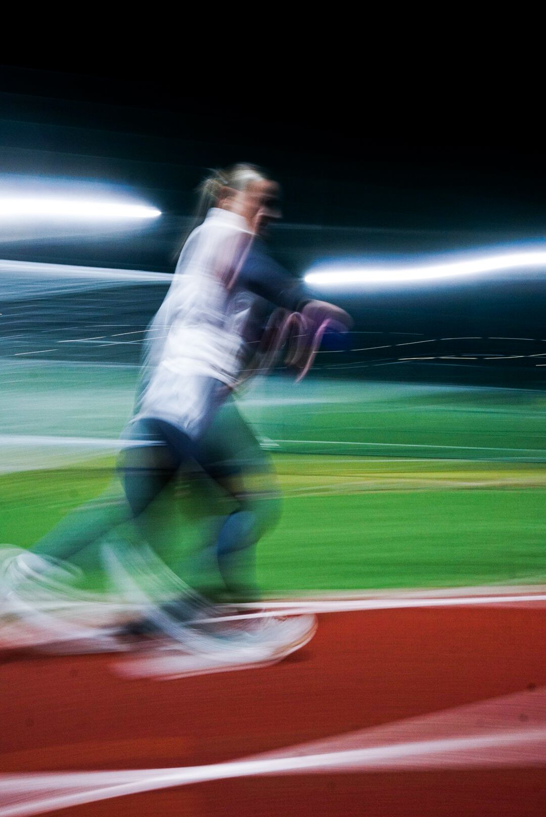 Motion blurred image of an athlete running on a red track at night with stadium lights in background.