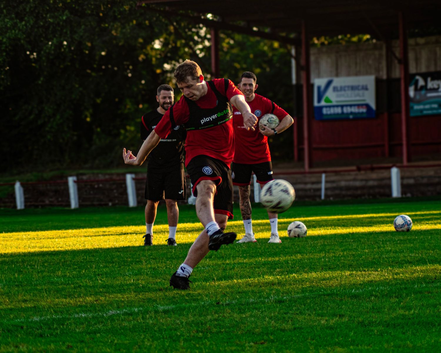 A soccer player in red kicks a ball during an outdoor training session on a green grass field.