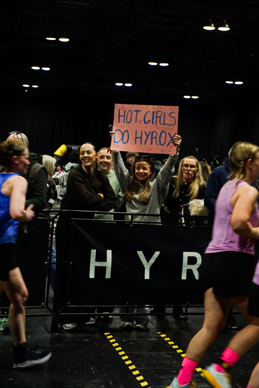 Spectators hold a Hot Girls Do HYROX sign while cheering at a HYROX fitness competition.