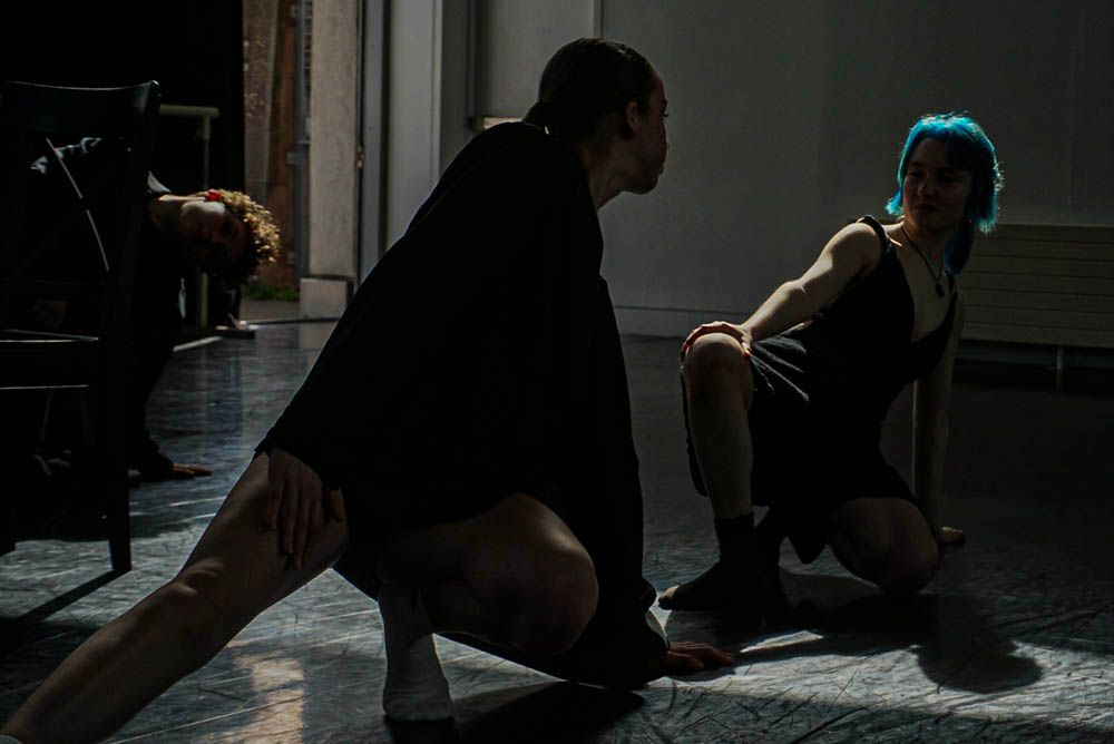 Two dancers in dramatic low-lit studio pose on floor in dark costumes during contemporary dance rehearsal.