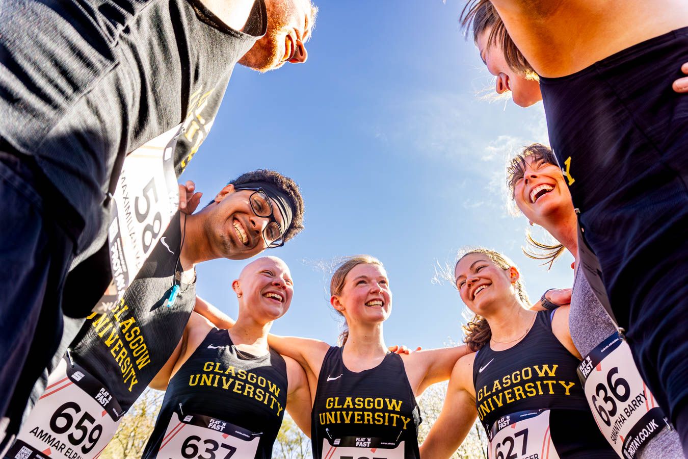 Glasgow University cross country running team celebrates together in a joyful huddle wearing numbered race bibs.