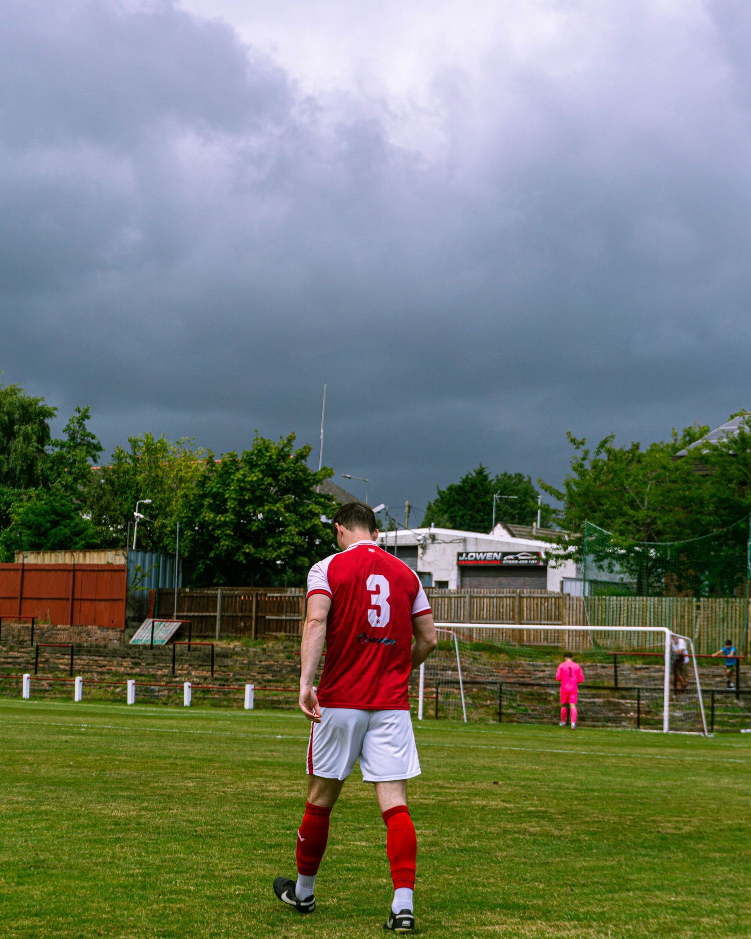Player number 3 in red and white stands on the pitch under dramatic storm clouds.