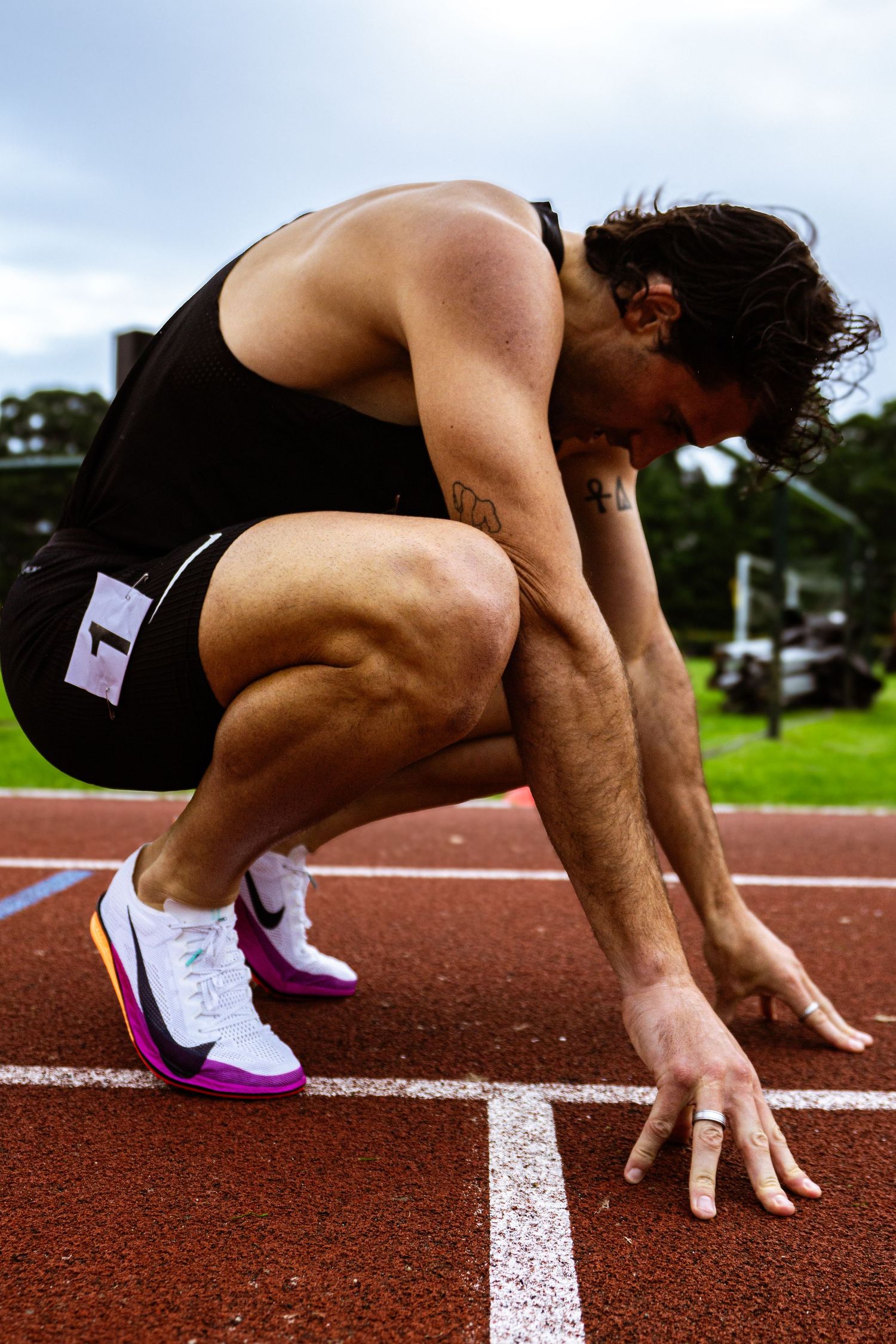 Male sprinter in black athletic wear crouches in starting position on red running track, wearing colorful Nike racing spikes.