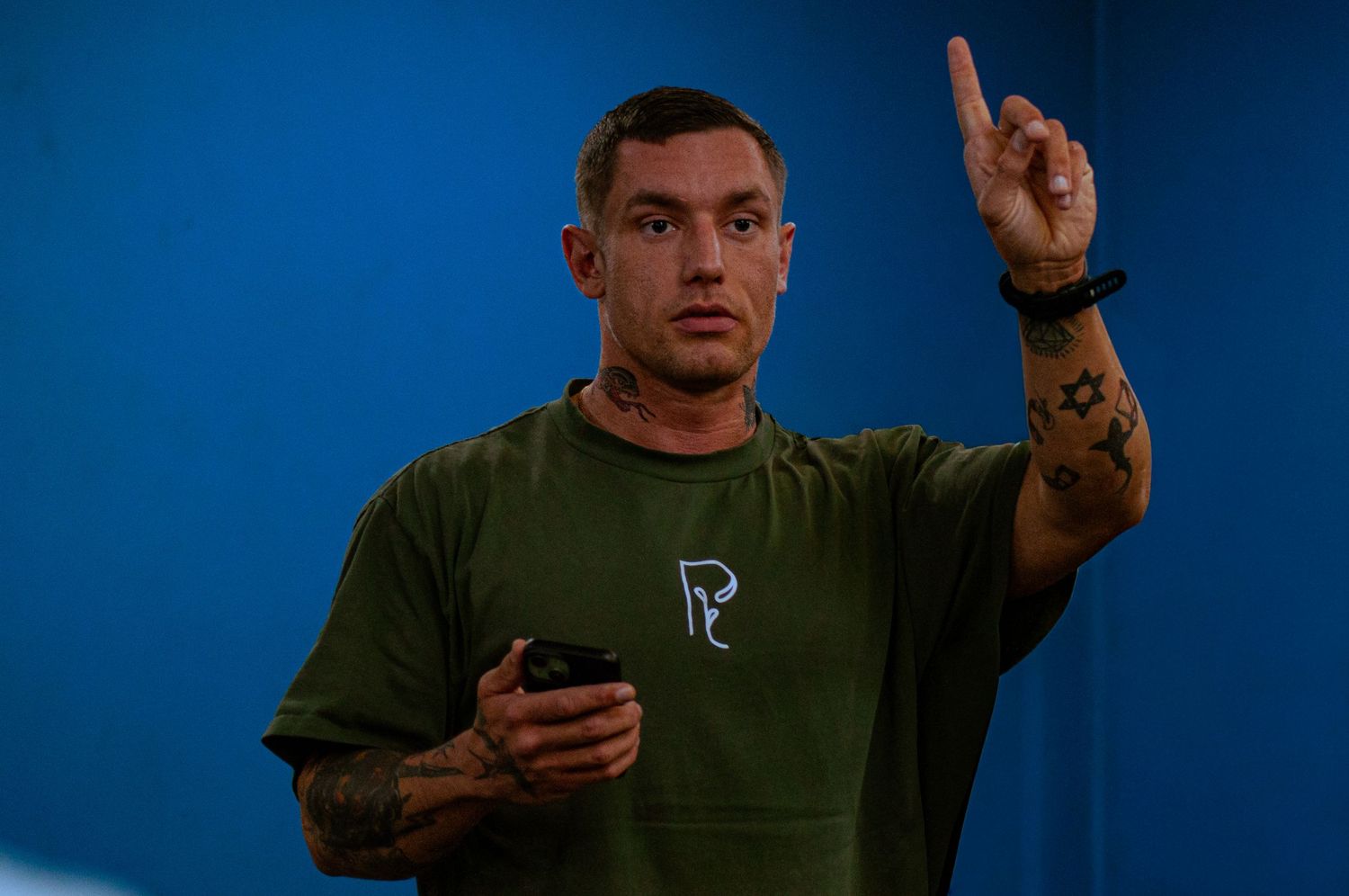 Man in green t-shirt holding phone and pointing finger up against blue background with tattoos visible.