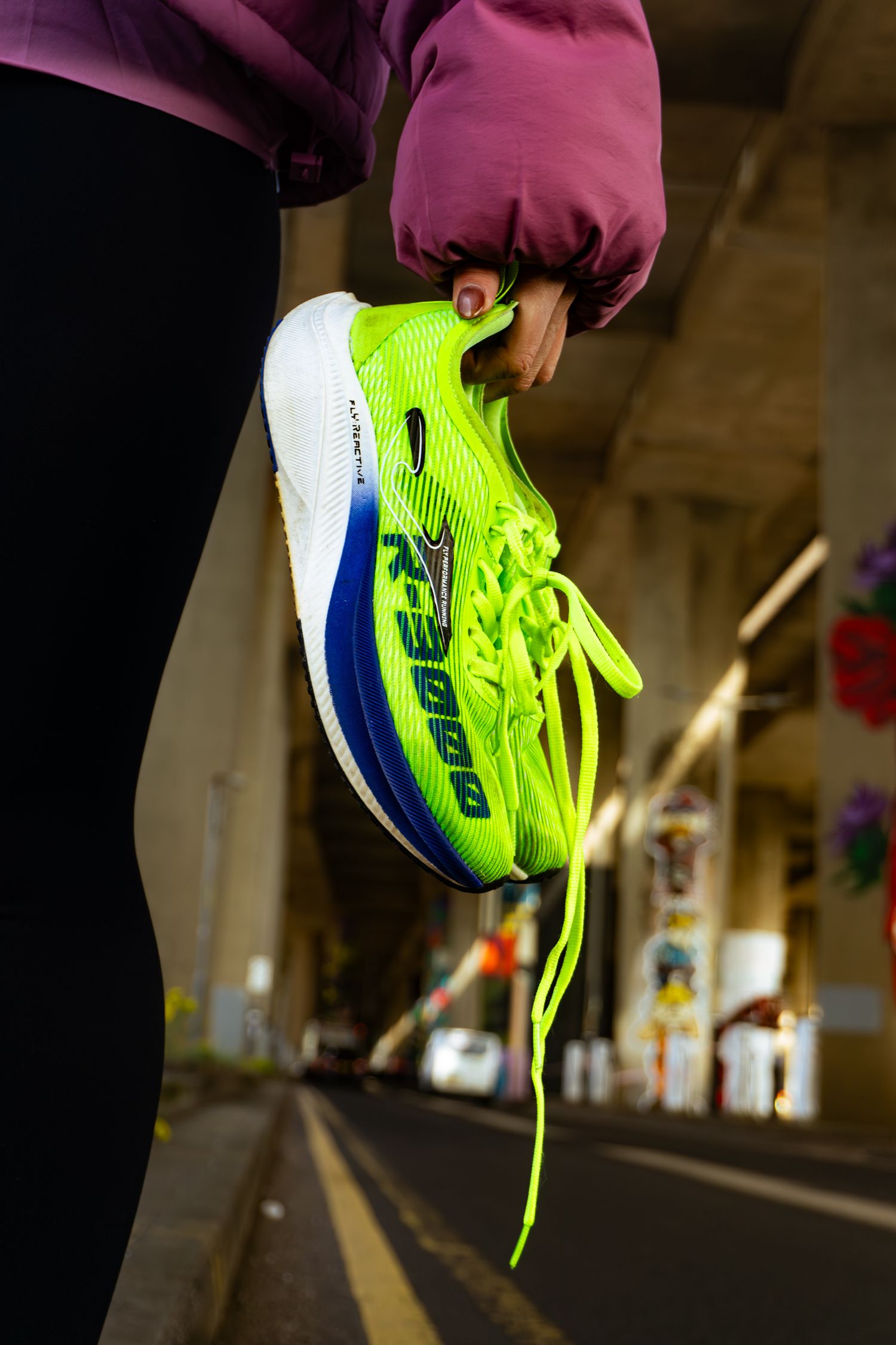 Hand holding neon green Joma R-3000 running shoe under urban bridge with blurred background.