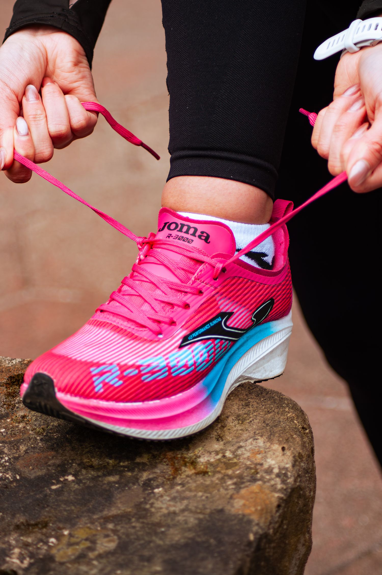 A woman ties the bright pink laces on her Joma R-3000 running shoes while seated on a stone surface.