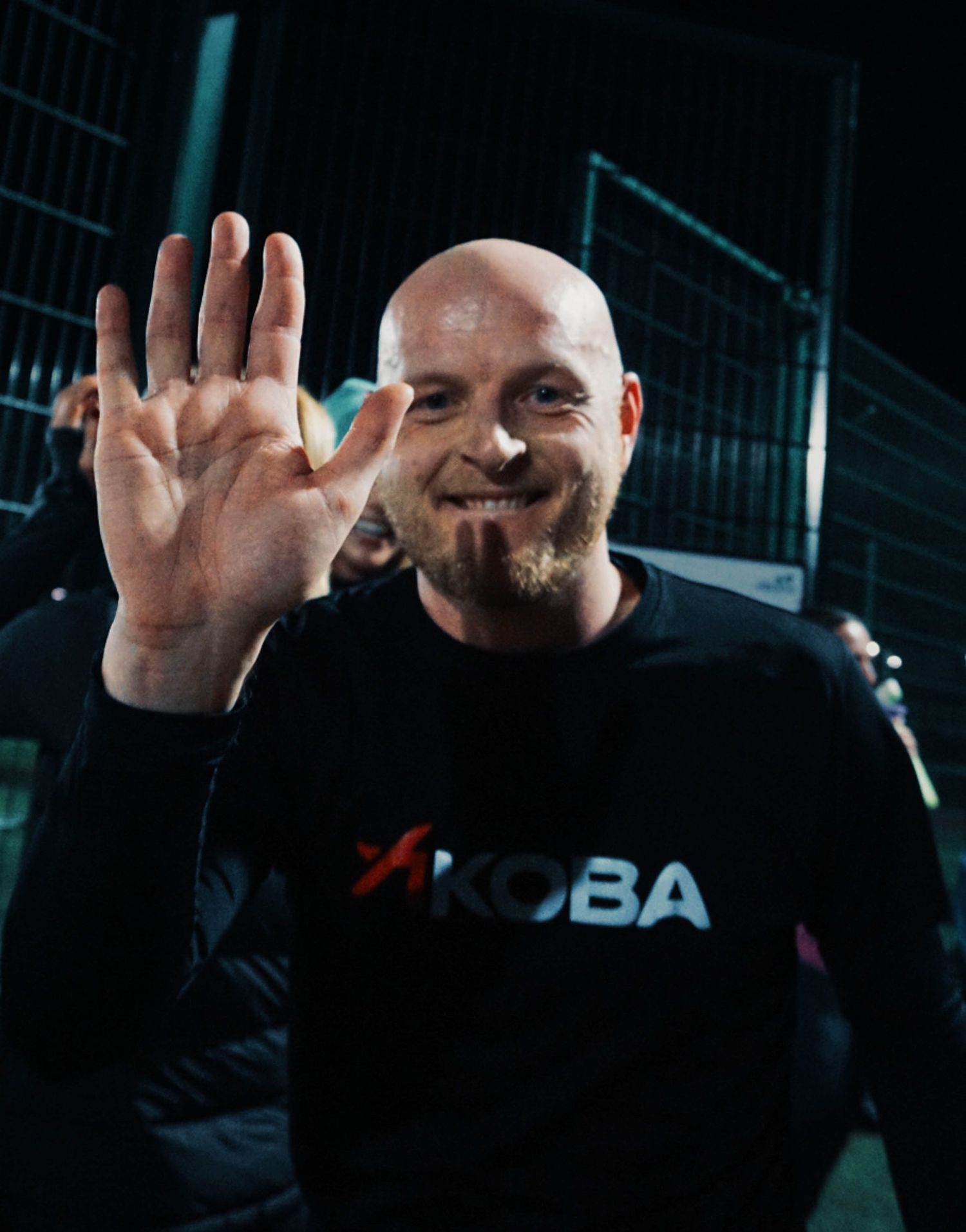 Bald man in black KOBA sweatshirt waves and smiles at the camera outdoors at night.