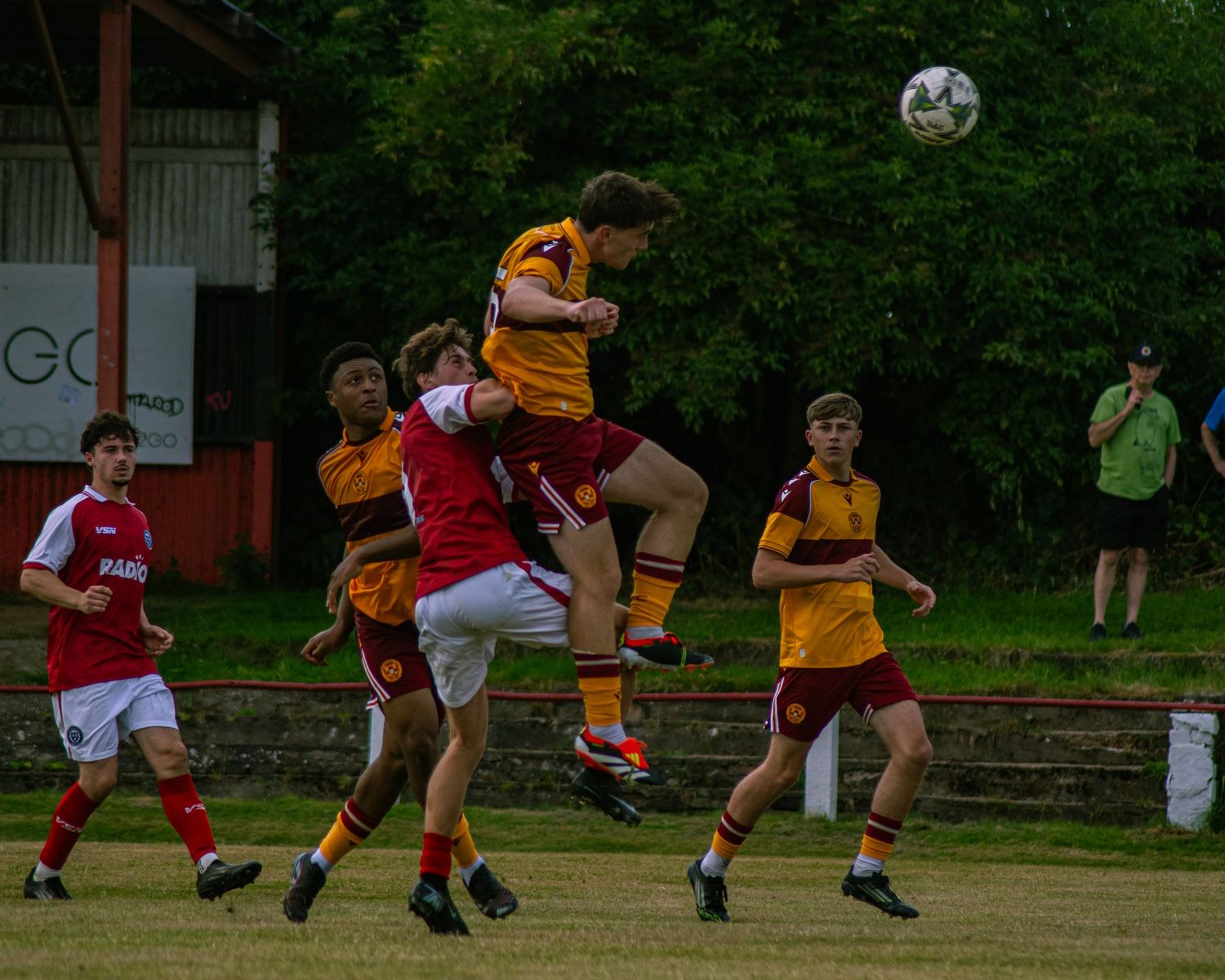 Youth soccer players compete for a header during an intense match, with teammates watching on a green field.