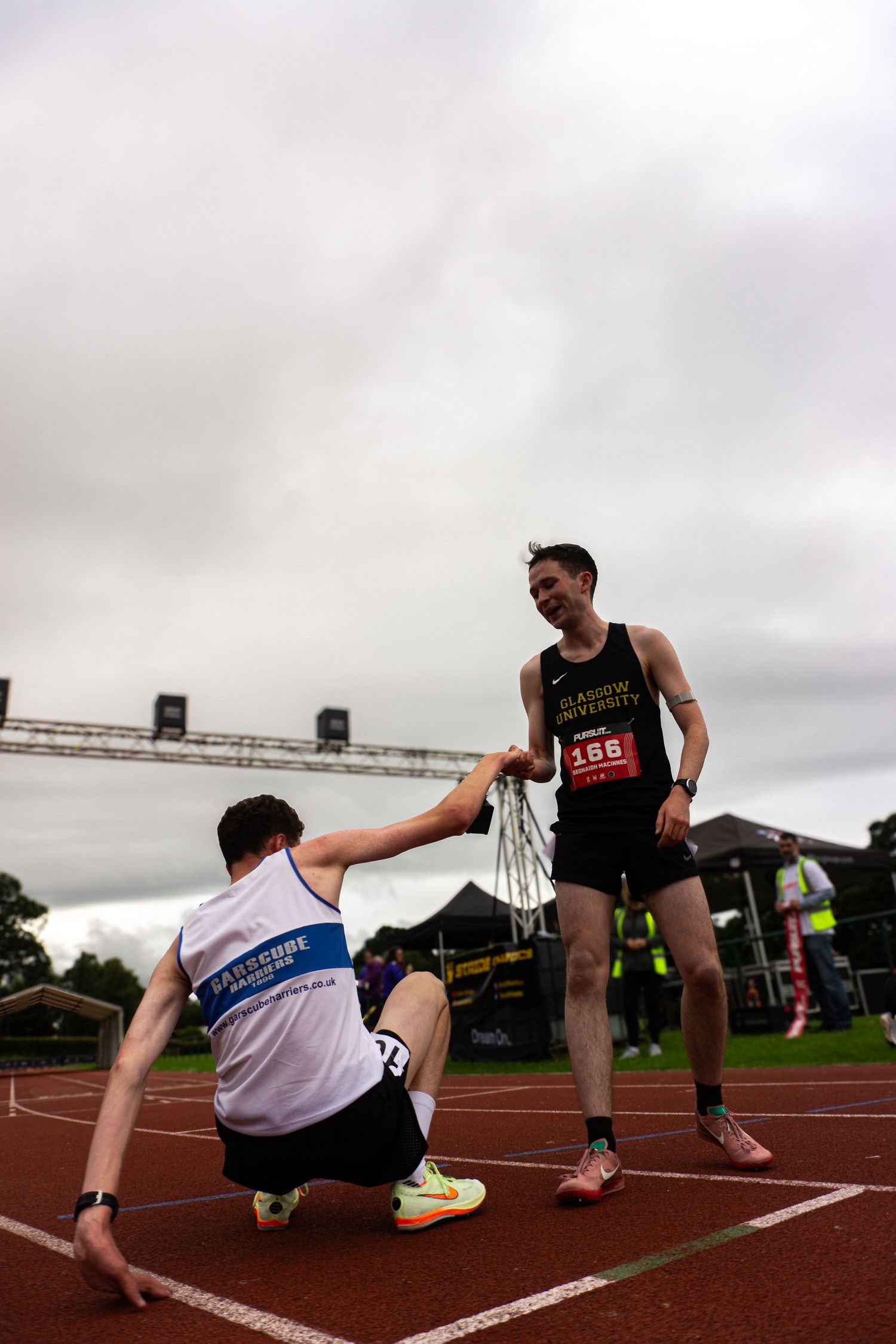 Runner 156 helps exhausted athlete rise from track at outdoor athletics competition on overcast day.