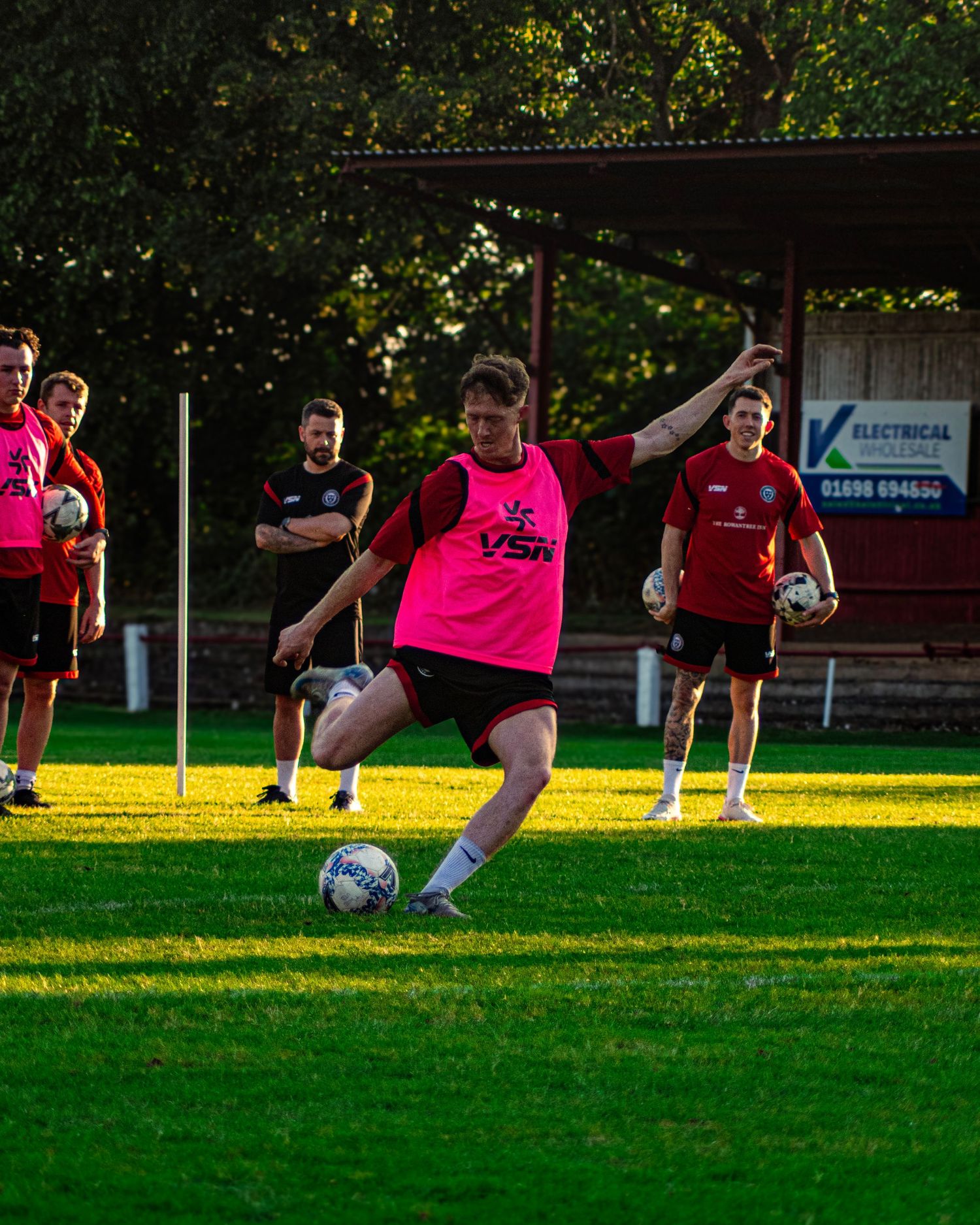 A footballer in a pink bib kicks a ball during training on a green pitch with teammates watching nearby.