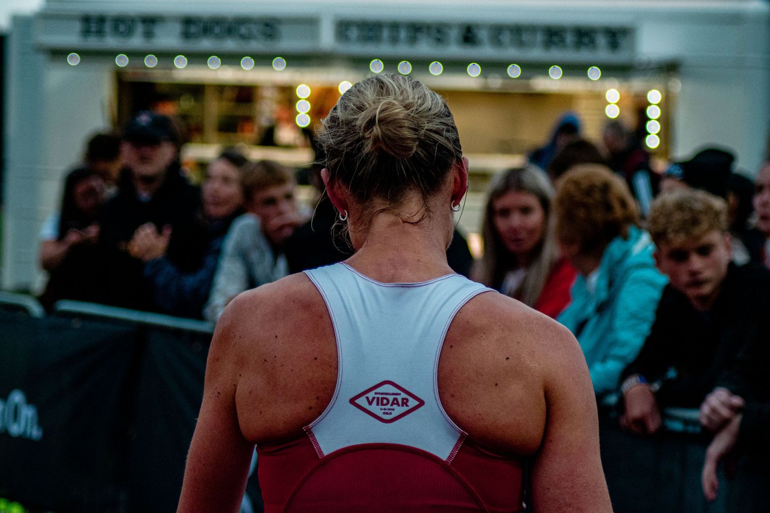 Female triathlete with VDAS logo on back of racing vest stands before crowd at outdoor food stall event.