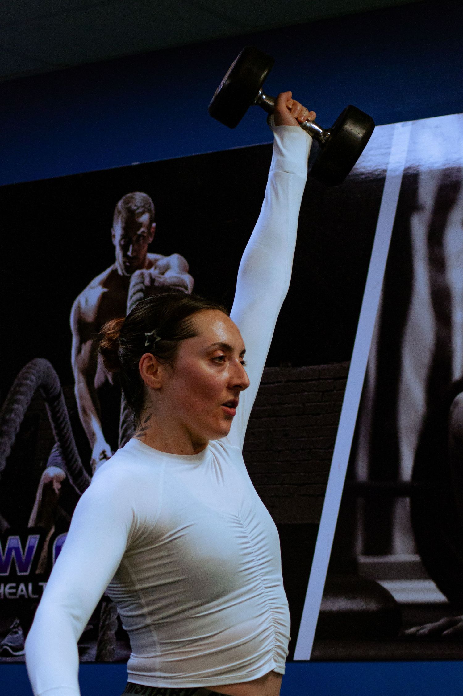 Athletic woman in white top lifting dumbbell overhead in gym.