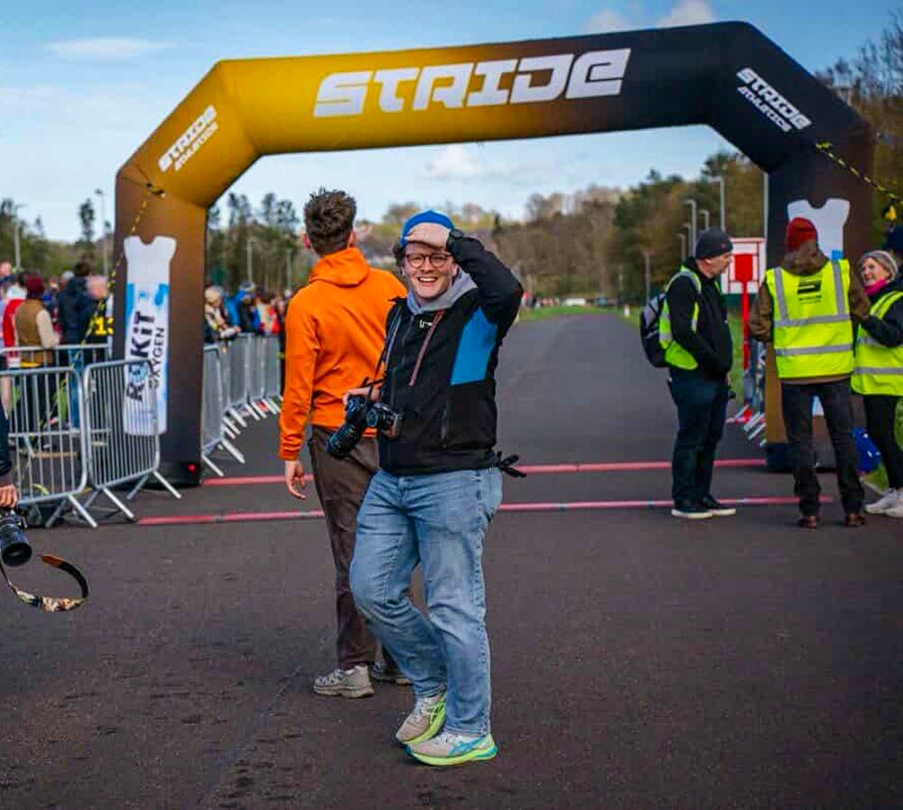 Smiling photographer stands under a Stride race finish line arch at an outdoor event.