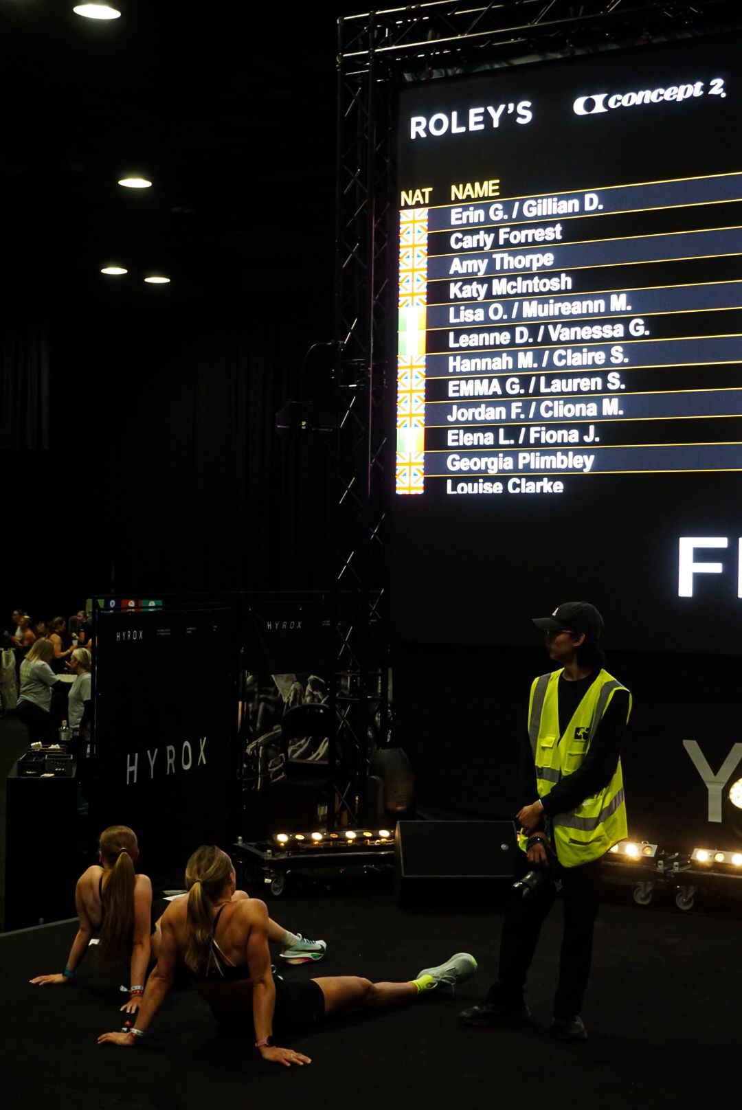 Athletes recover on the floor beneath a HYROX leaderboard displaying competitor names and rankings.