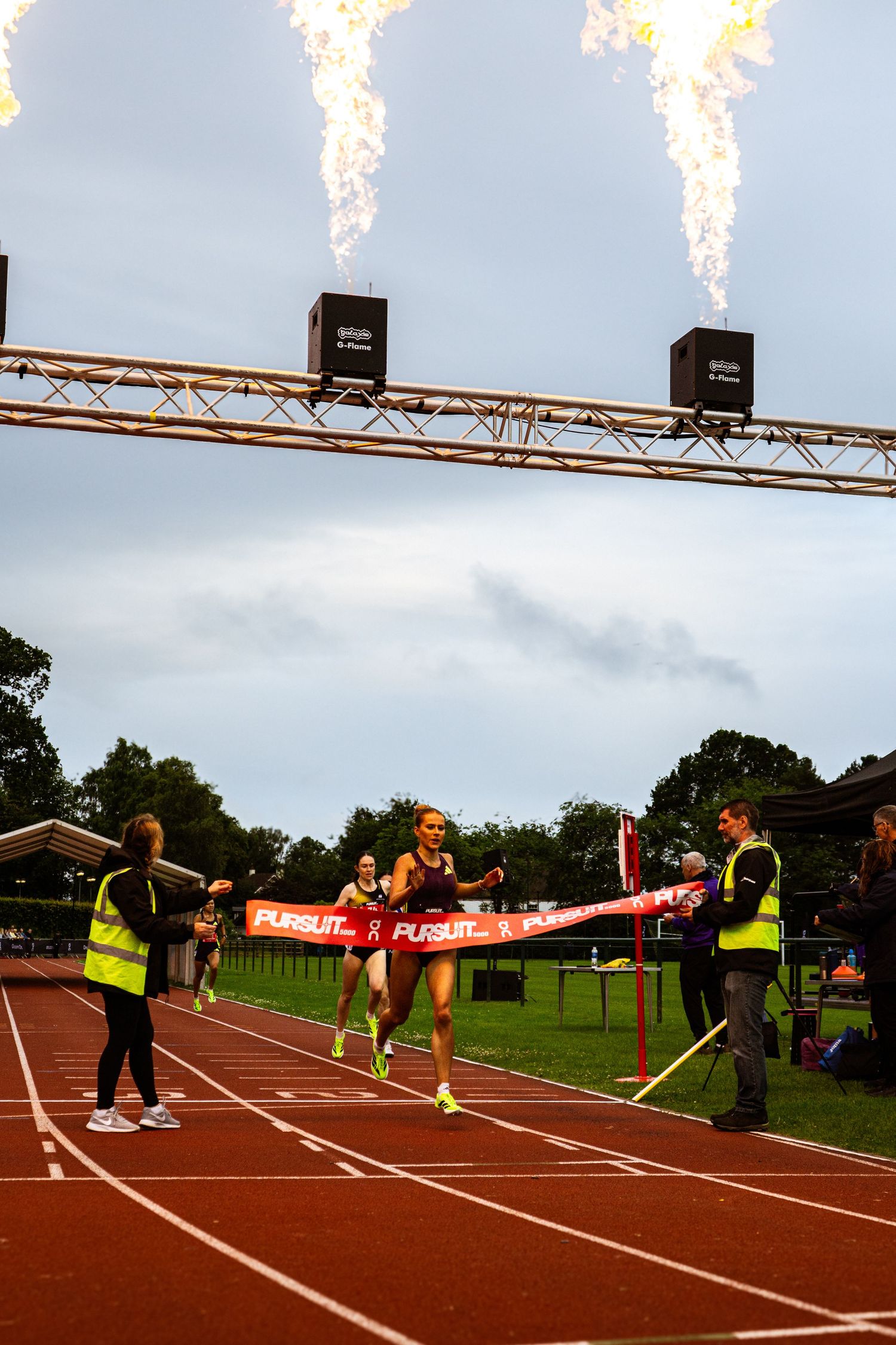 Female runner breaks finish line tape at track event with dramatic fire cannons overhead on cloudy day.