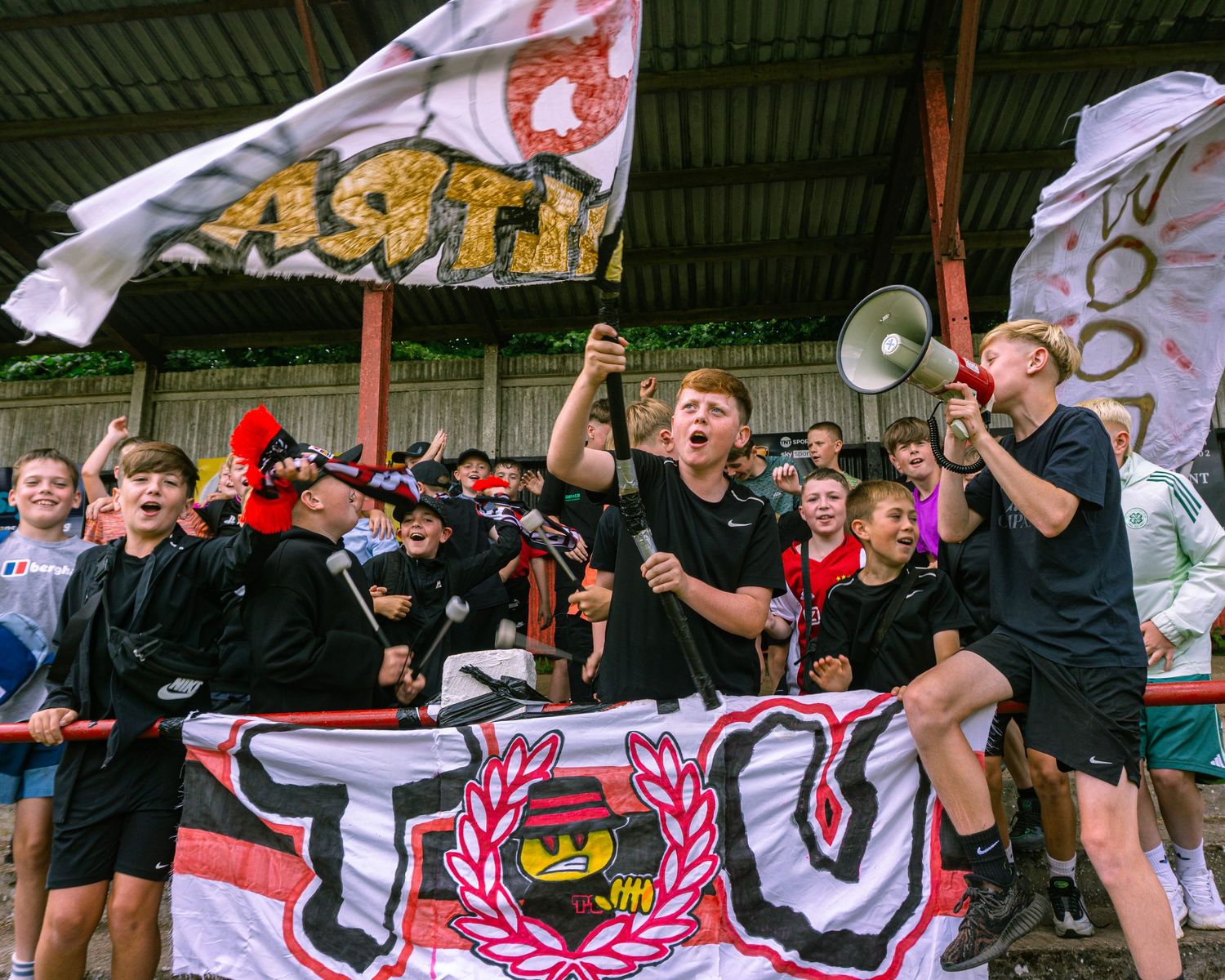 Energetic football fans cheering with flags, scarves, and a megaphone in stadium stands during a match.