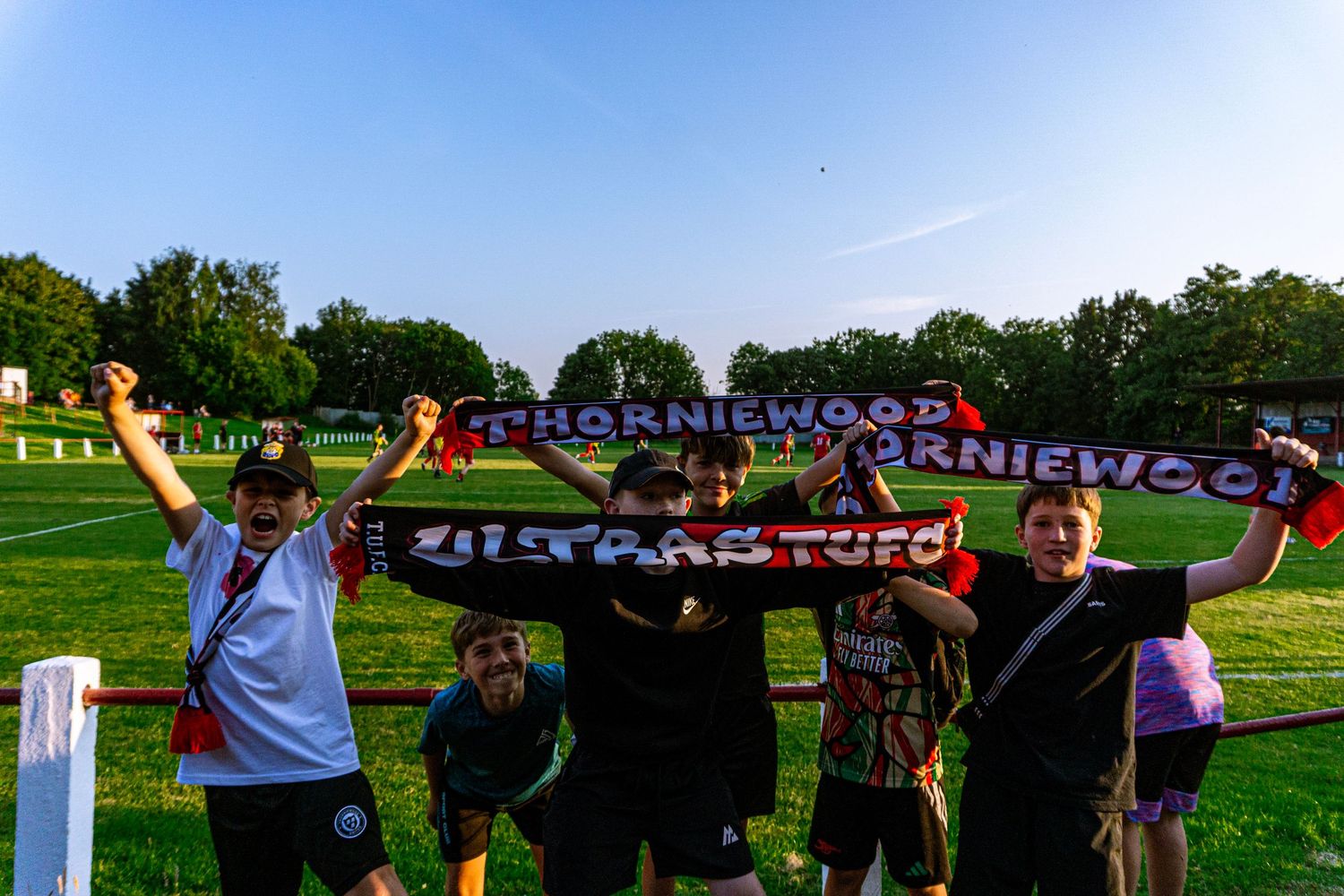 Fans holding Thorniewood FC scarves aloft at an outdoor football ground.