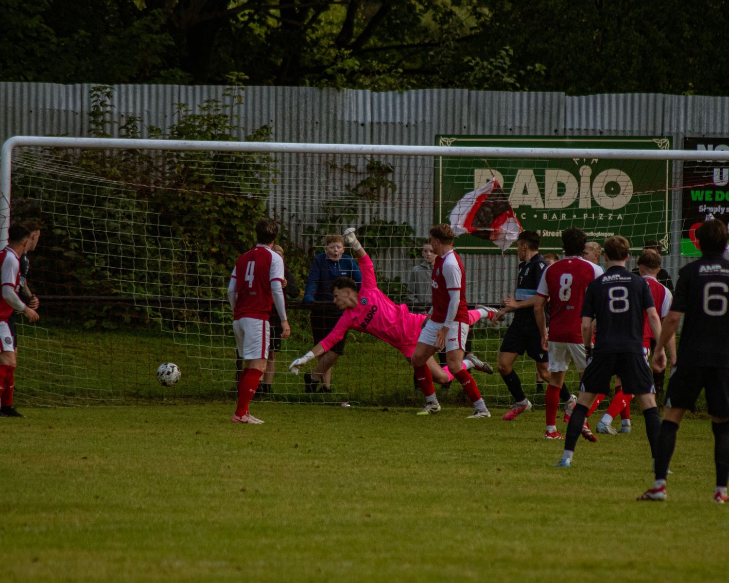A goalkeeper in pink dives to make a save during a soccer match with players in red and black uniforms on a green field.