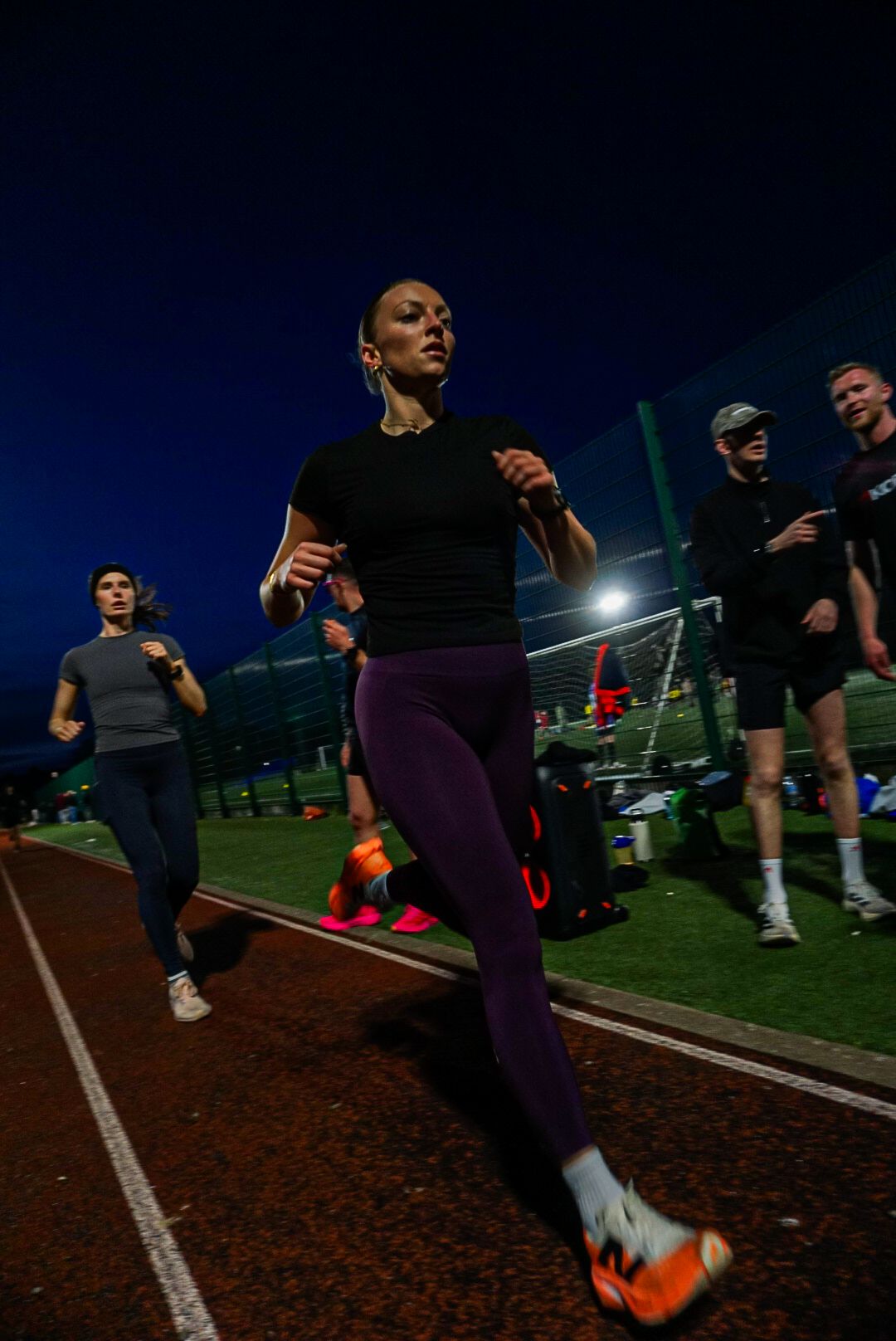 Female athlete in black top and purple leggings running on a track during a night session.