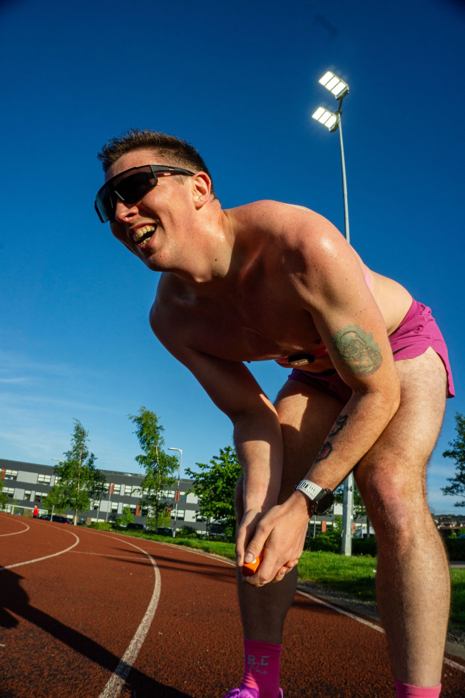 Shirtless male athlete in pink shorts laughing on a red running track under a clear blue sky.