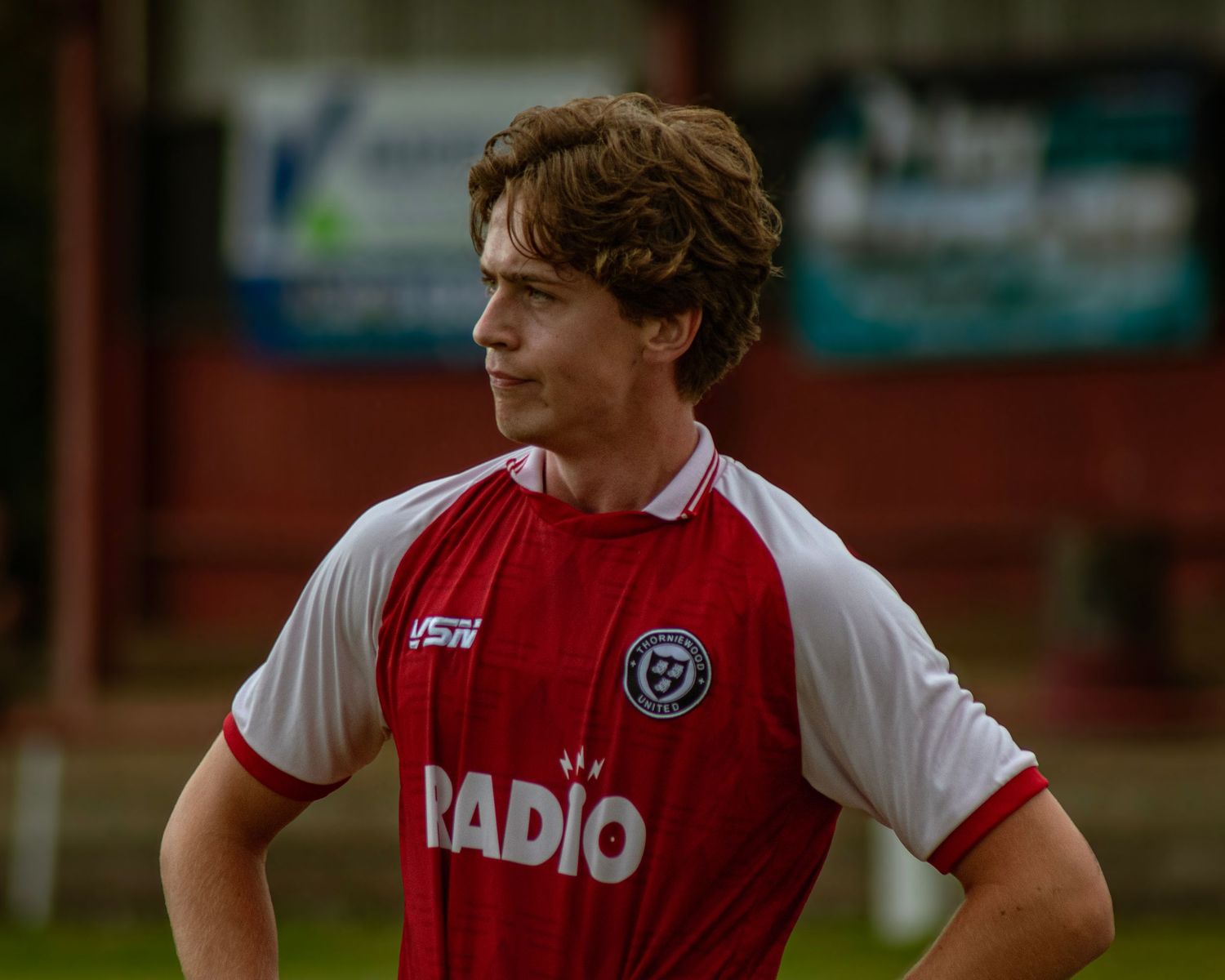A curly-haired footballer in a red and white Radio jersey looks sideways during a match.