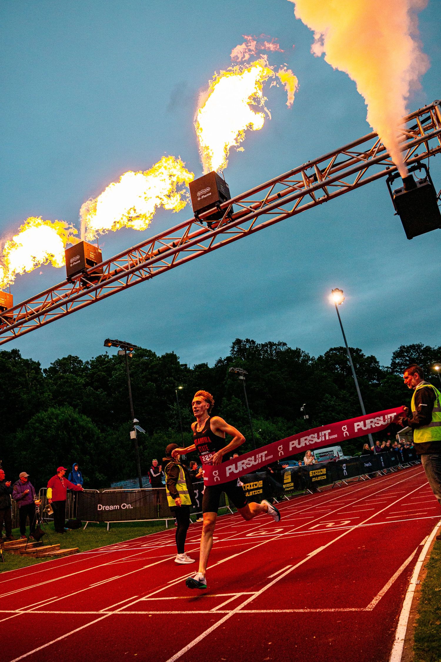 Athletes sprint past a dramatic fire display at the finish line of a Pursuit race on a red track at dusk.