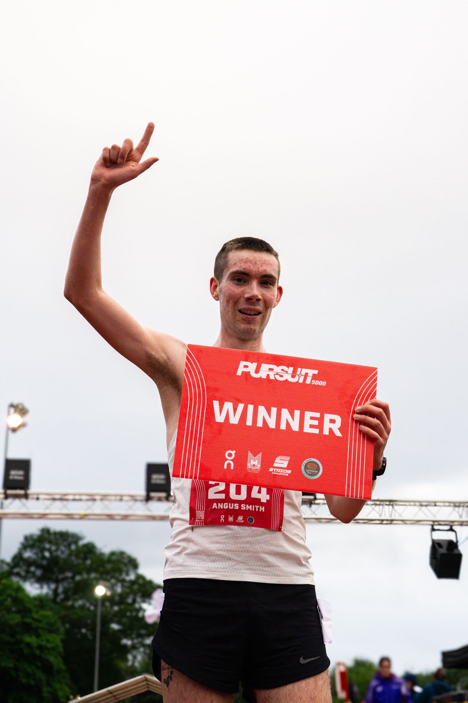 Winner Angus Smith celebrates victory at a Pursuit running event, holding a red winner sign and raising his hand.