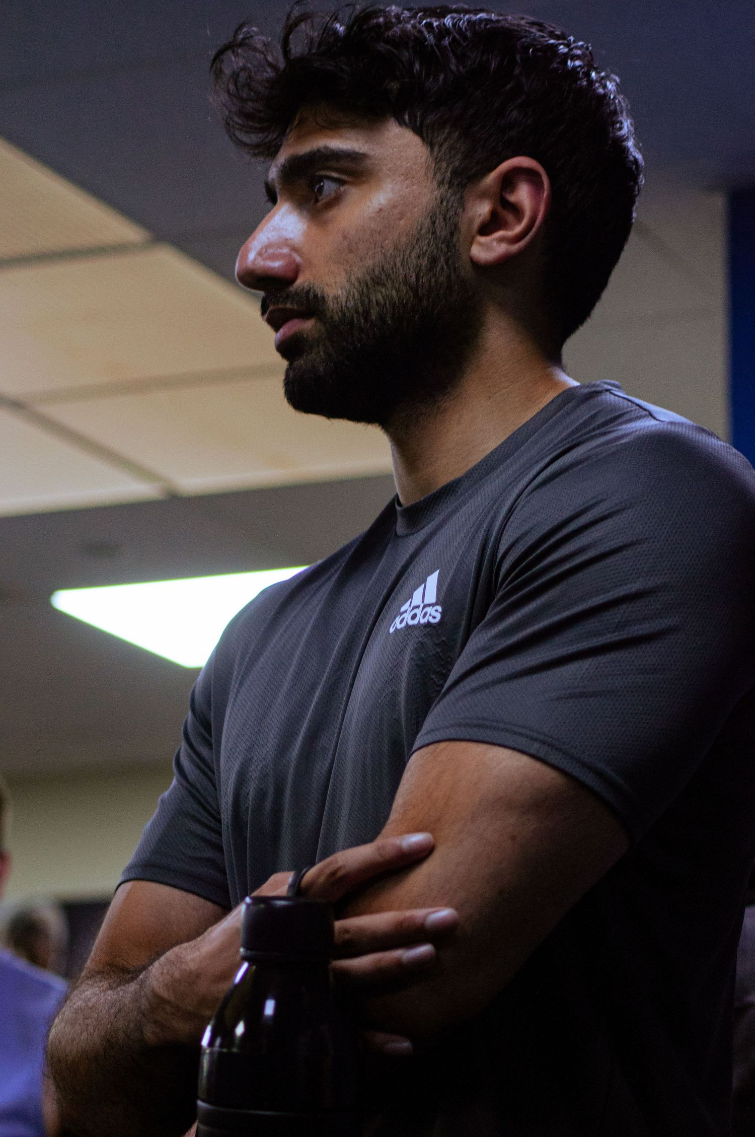 Muscular man in an Adidas shirt standing with arms crossed holding a water bottle in a gym.