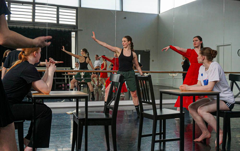 Ballet students rehearse at the barre while instructor in red dress demonstrates graceful arm positions.