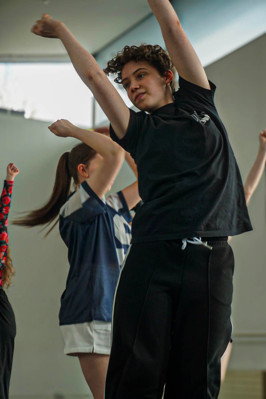 A curly-haired dancer in a black outfit leaps with arms raised during a dynamic dance class in a bright studio.