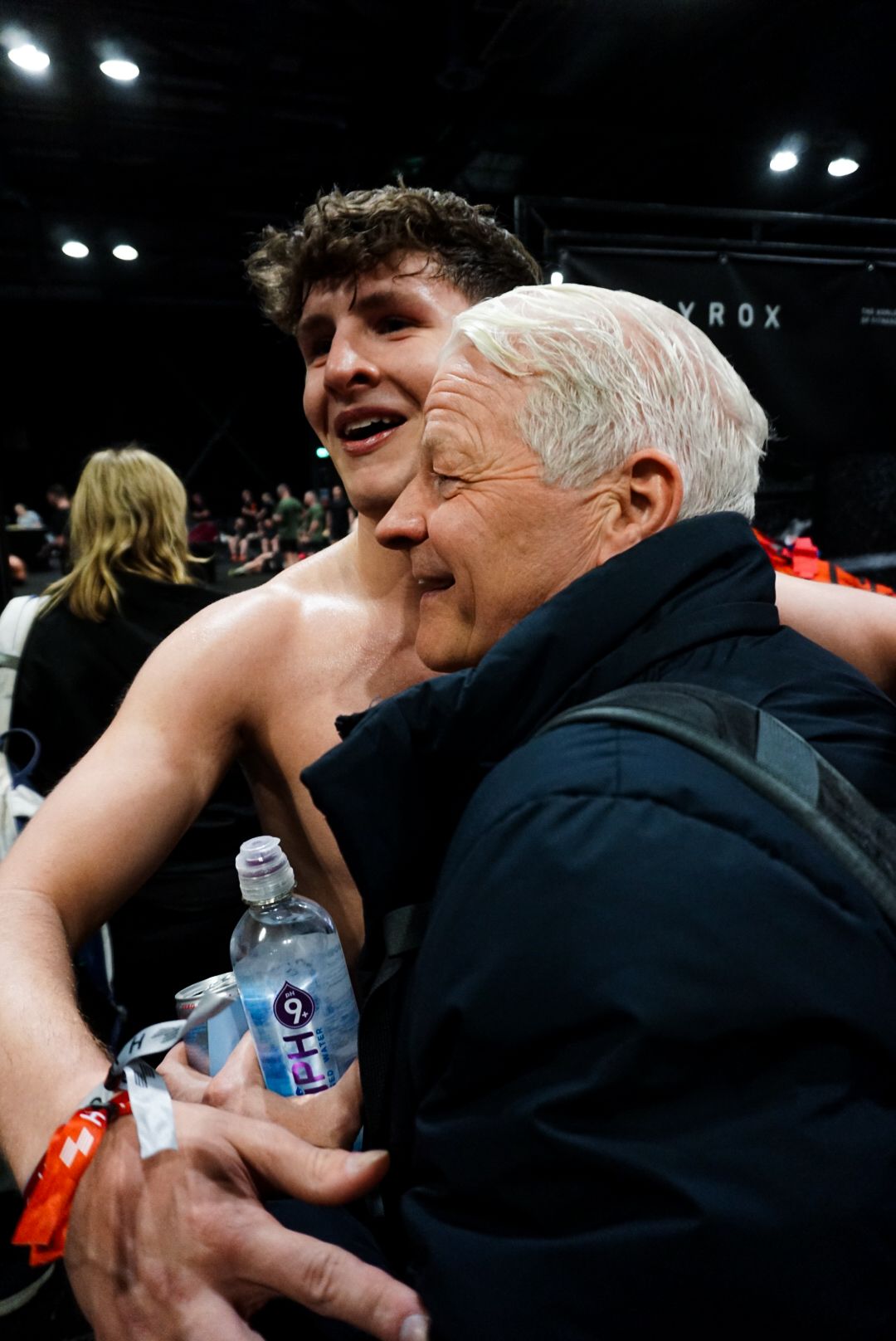 Young shirtless athlete embraces an older man after competing at a HYROX fitness event.