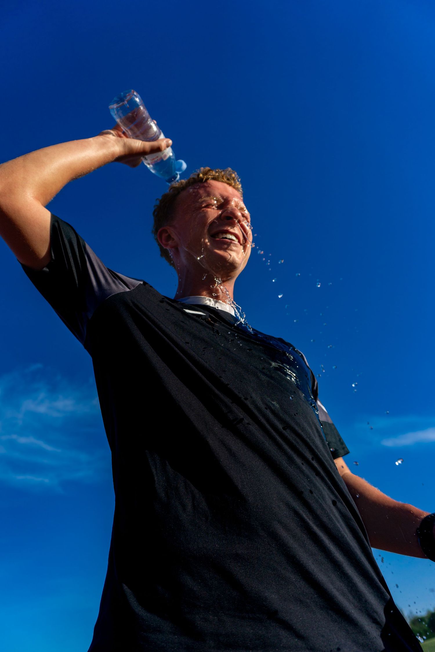 Male athlete pouring water over his head from a bottle, laughing under a bright blue sky after a workout.