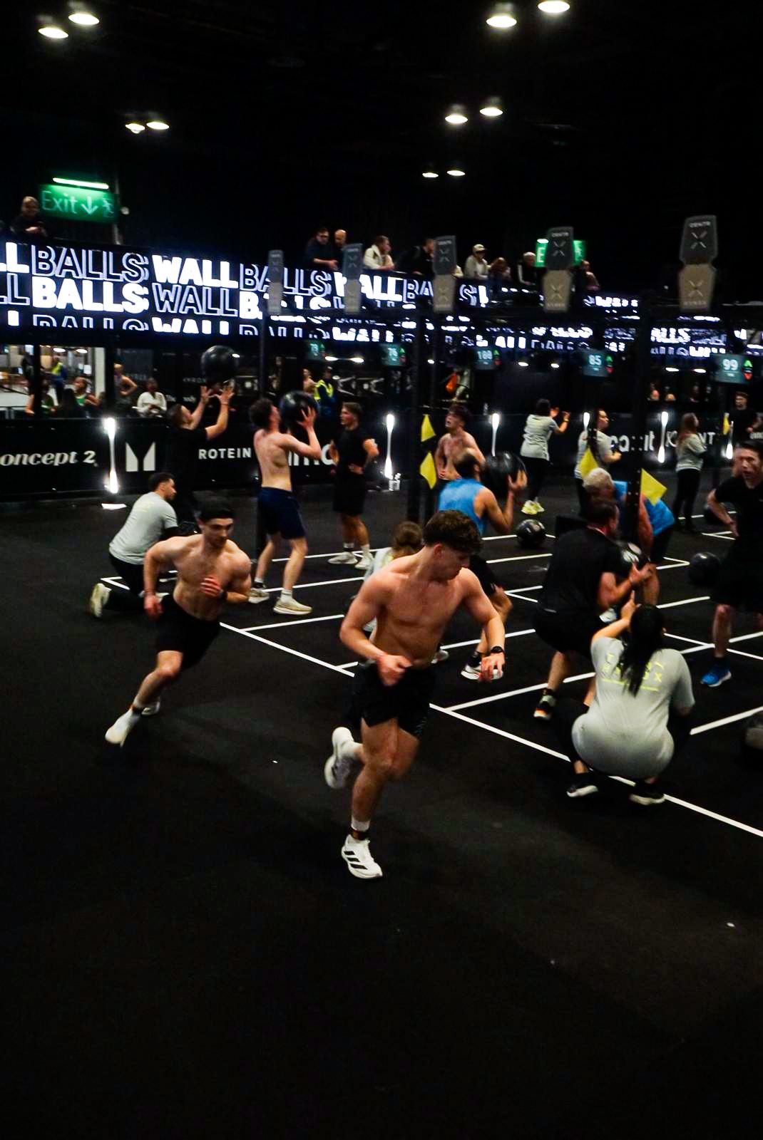 Large group of shirtless male athletes sprinting across a dark gym floor during a fitness event.