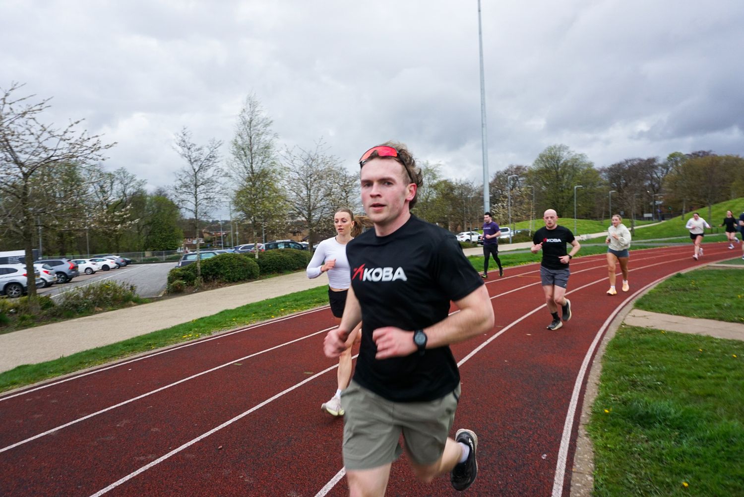 Group of runners on red athletics track led by man wearing red cap and Koba shirt.