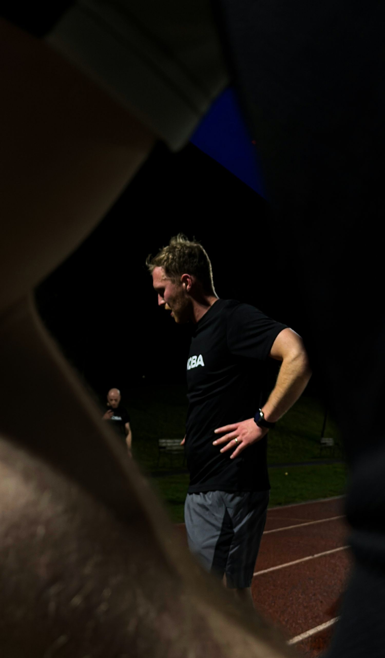 Athlete in black NBA shirt resting with hands on hips on a running track at night.