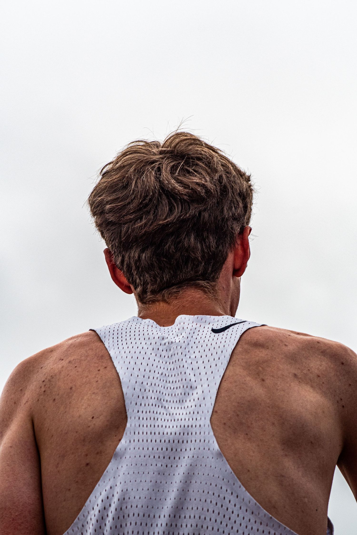 Back view of male runner in white Nike mesh singlet with sweat-dampened hair against grey sky.