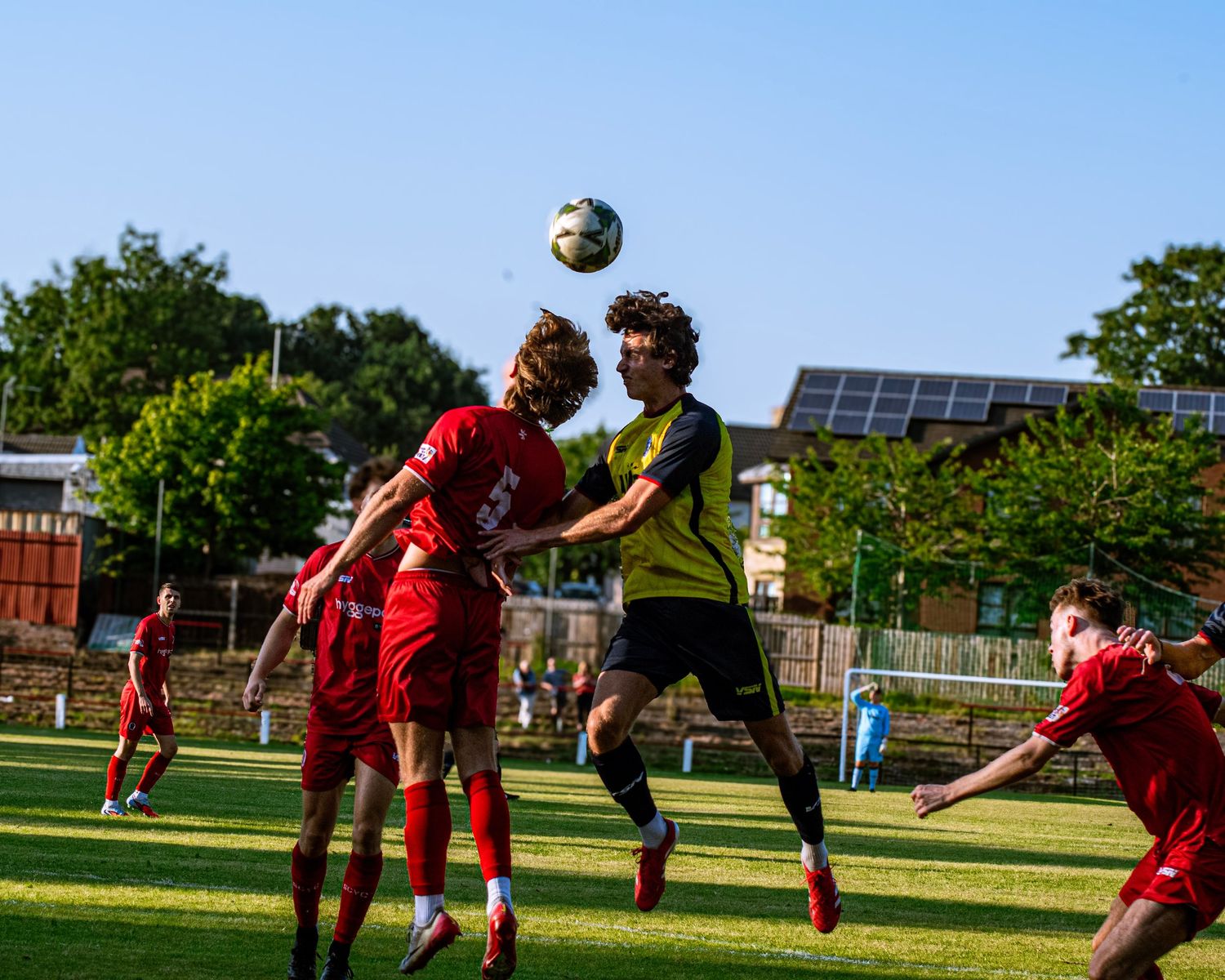 Soccer players leap to head the ball during an outdoor match on a sunny day, with red and yellow jerseys competing.