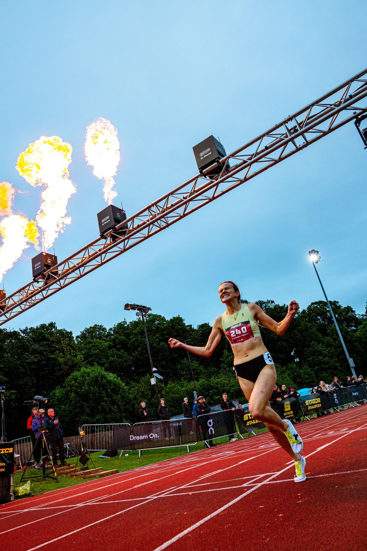 Female athlete crosses finish line on red track with dramatic fire pyrotechnics at night race event.