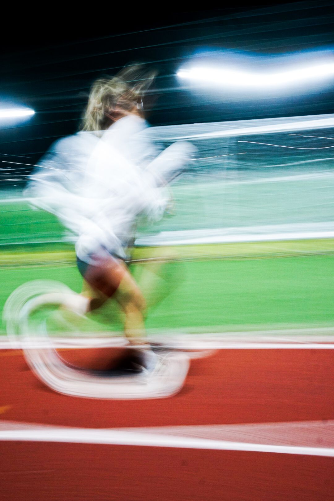 Long exposure motion blur image of a female runner with flowing hair sprinting on a red track at night.