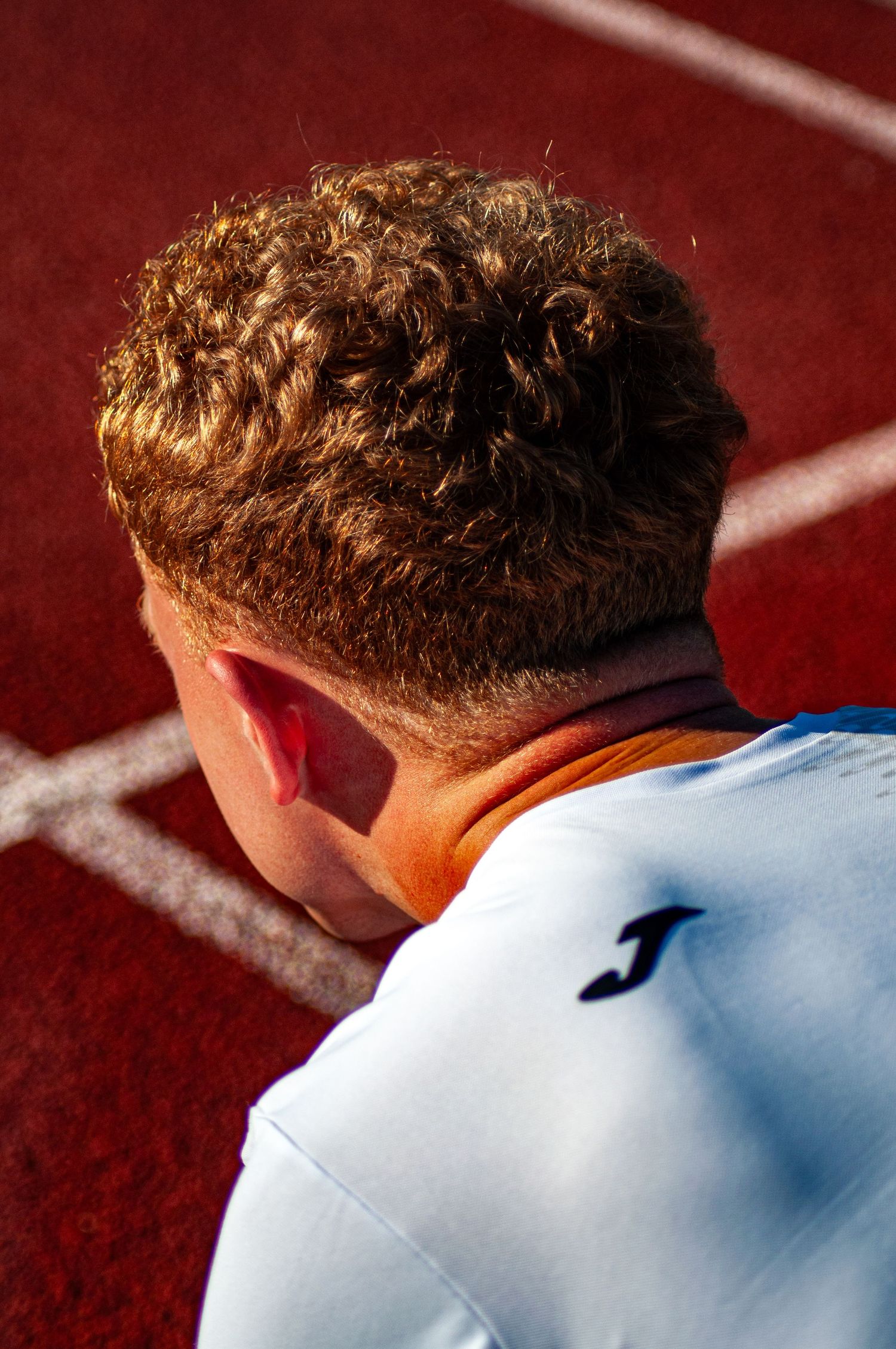 Close-up rear view of a male athlete with curly hair wearing a Joma jersey on a red running track.