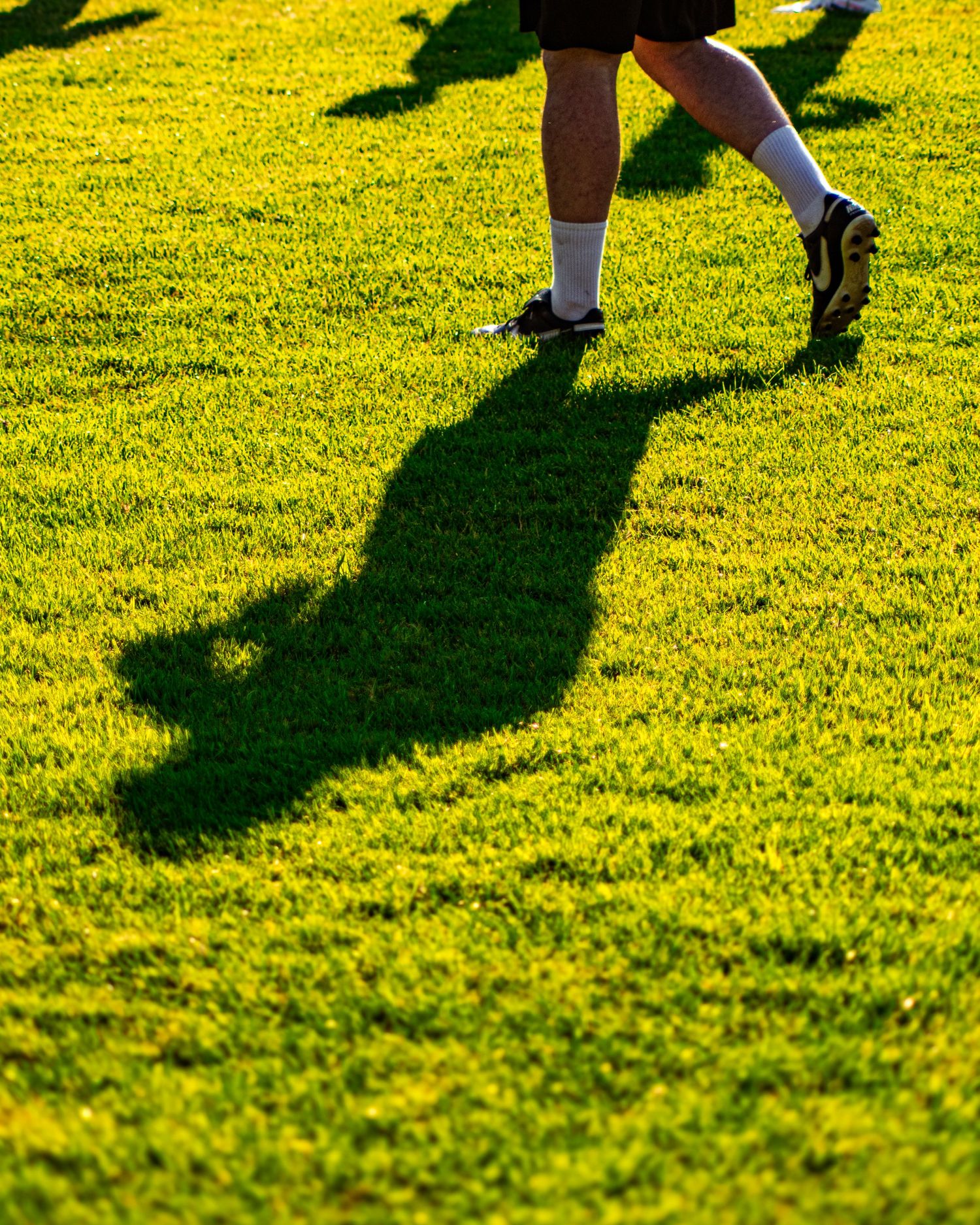 Person in cleats and white socks casting a long shadow while walking on a bright green grass field.