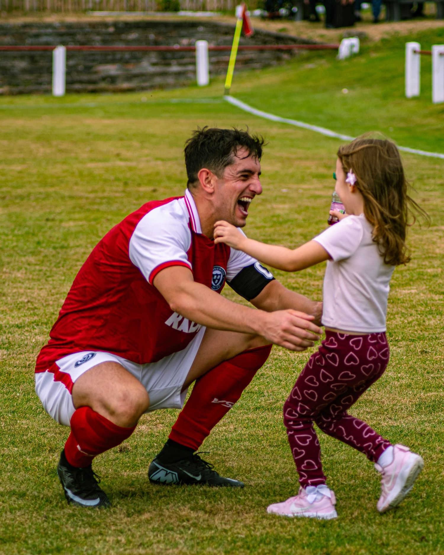 Soccer player in red kit crouches joyfully greeting a young girl on a grass pitch after a match.