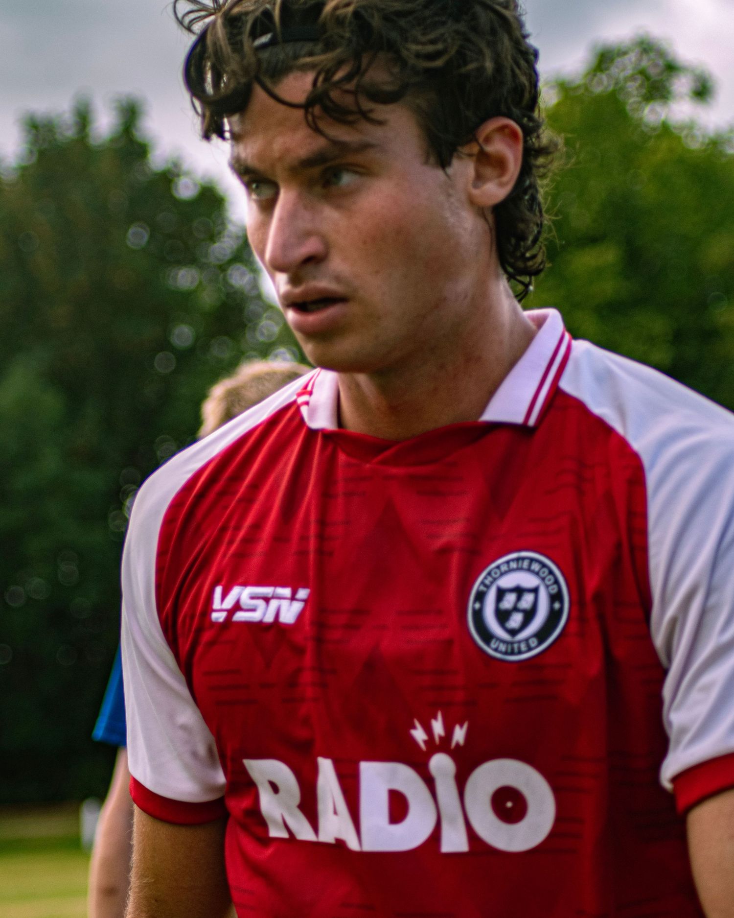 Young male soccer player in red and white VSN Radio jersey with curly hair on outdoor field.