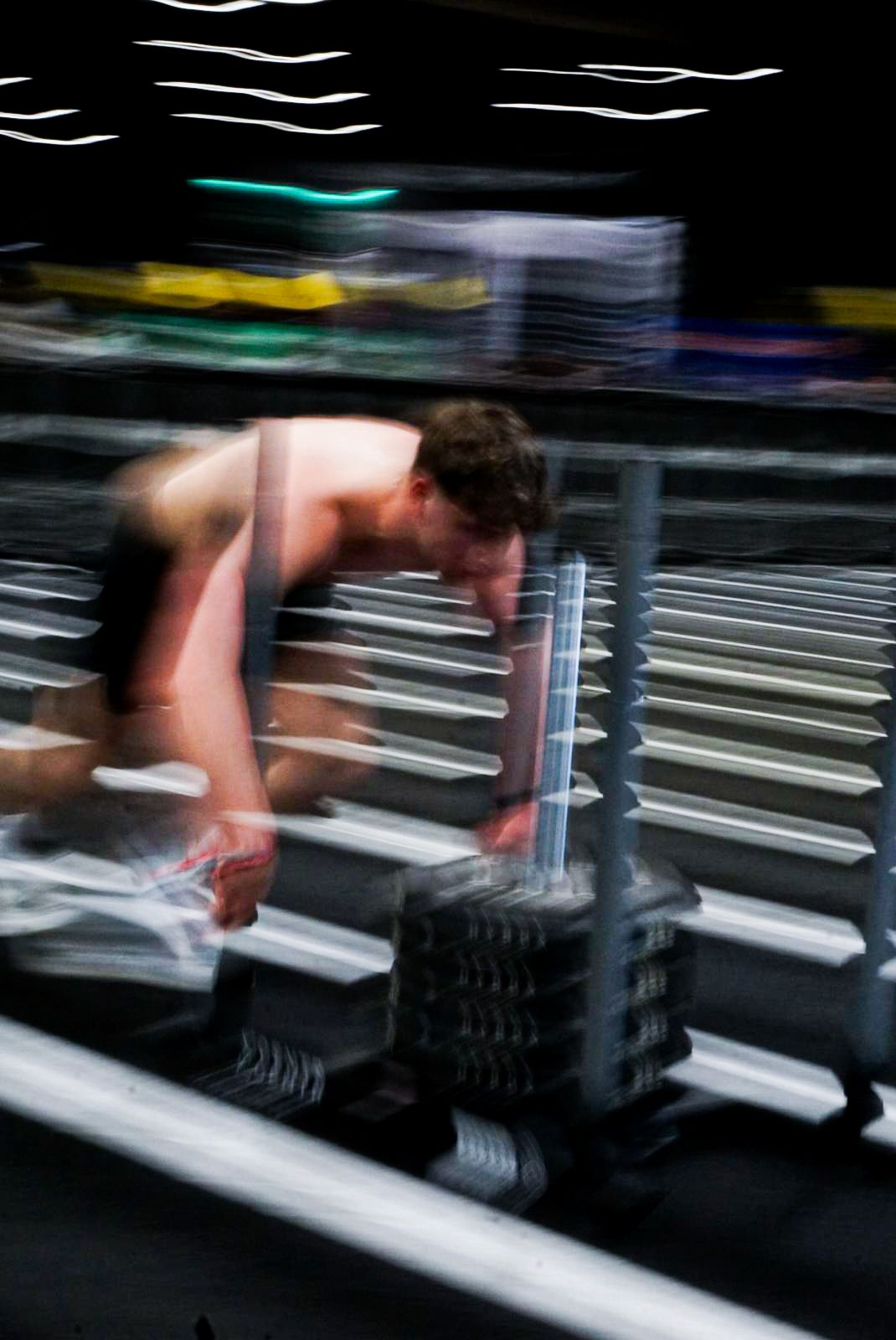Motion blur photo of shirtless athlete picking up dumbbells from a rack during a fitness event.