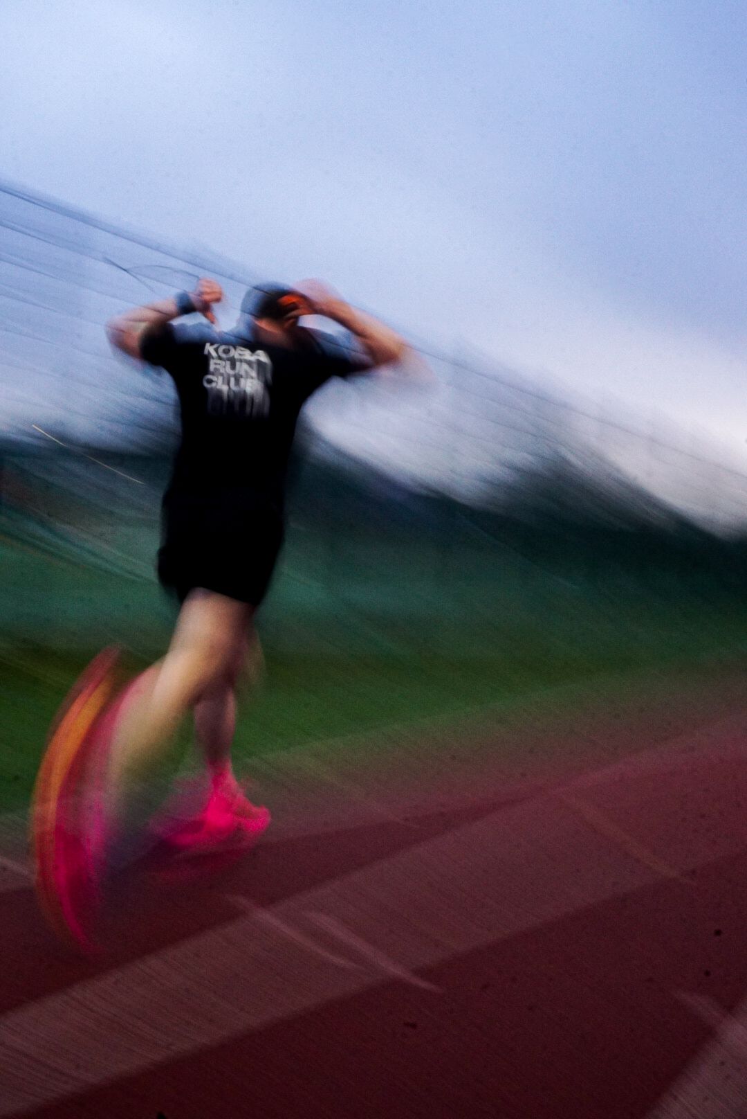 Motion blur action shot of runner in black Koba Run Club shirt on red track.