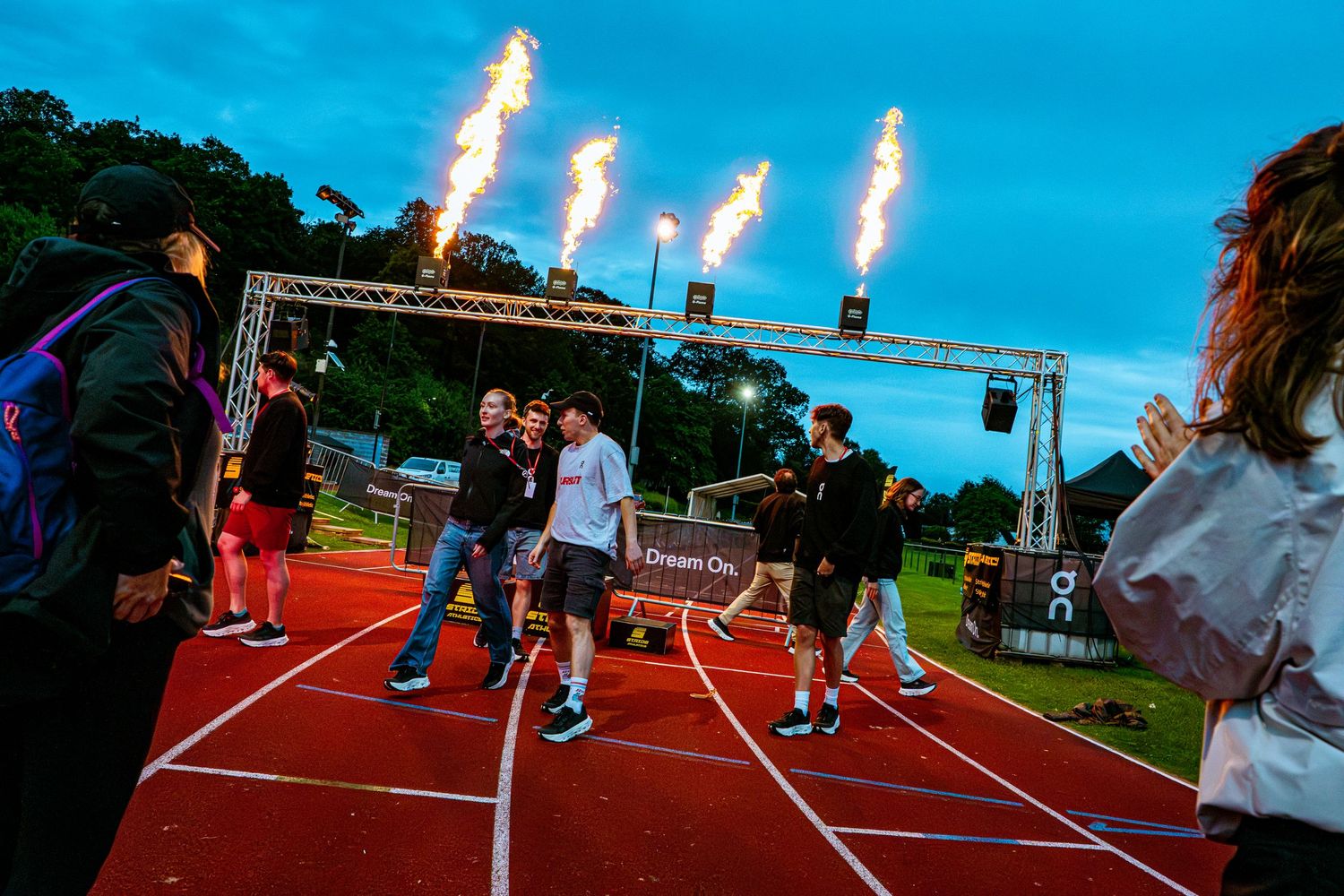Athletes walk on a red track under dramatic fire jets at dusk during an exciting outdoor sporting event.